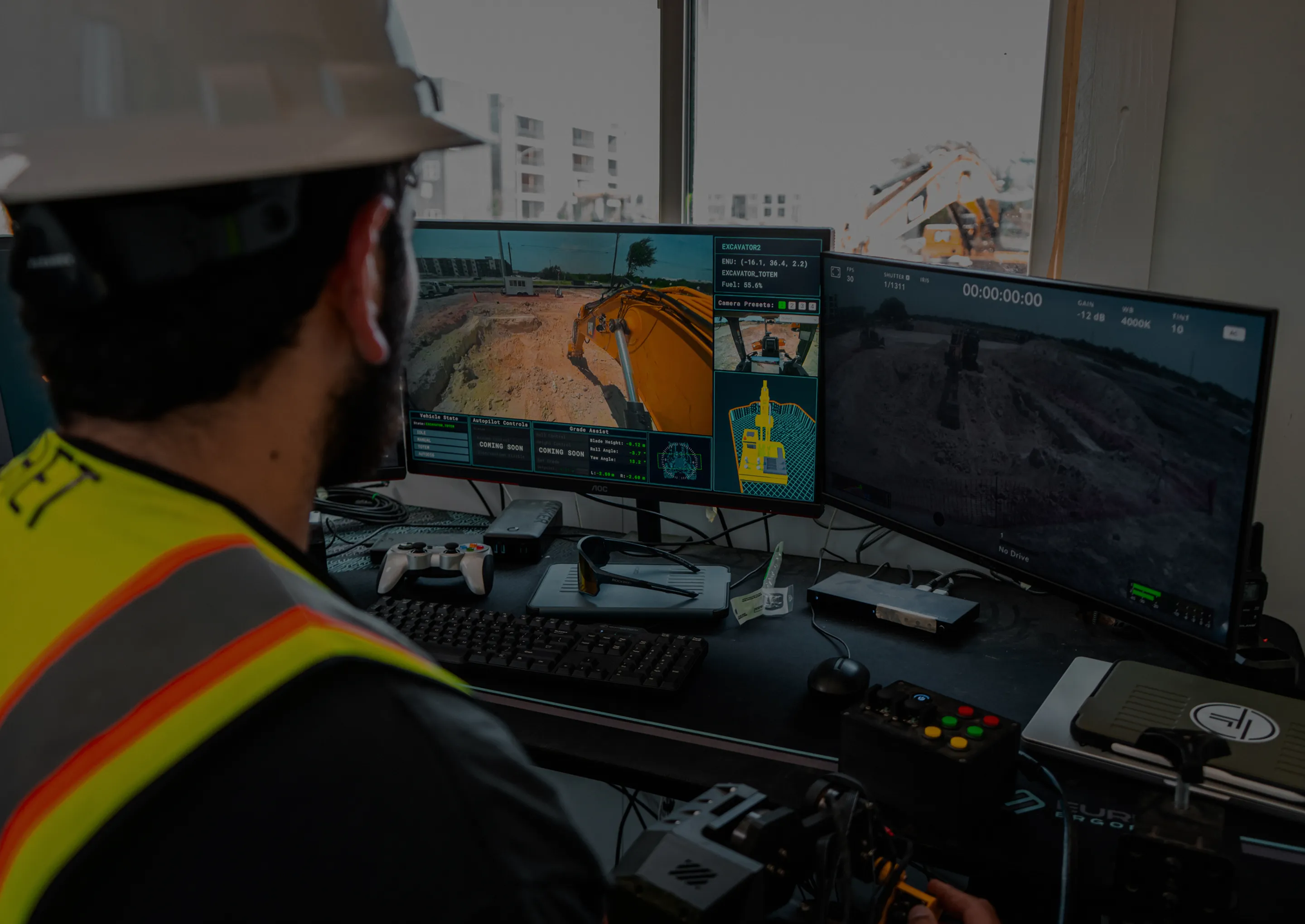 Construction worker operating heavy machinery remotely from a control station with dual monitors showing live excavator camera feeds.