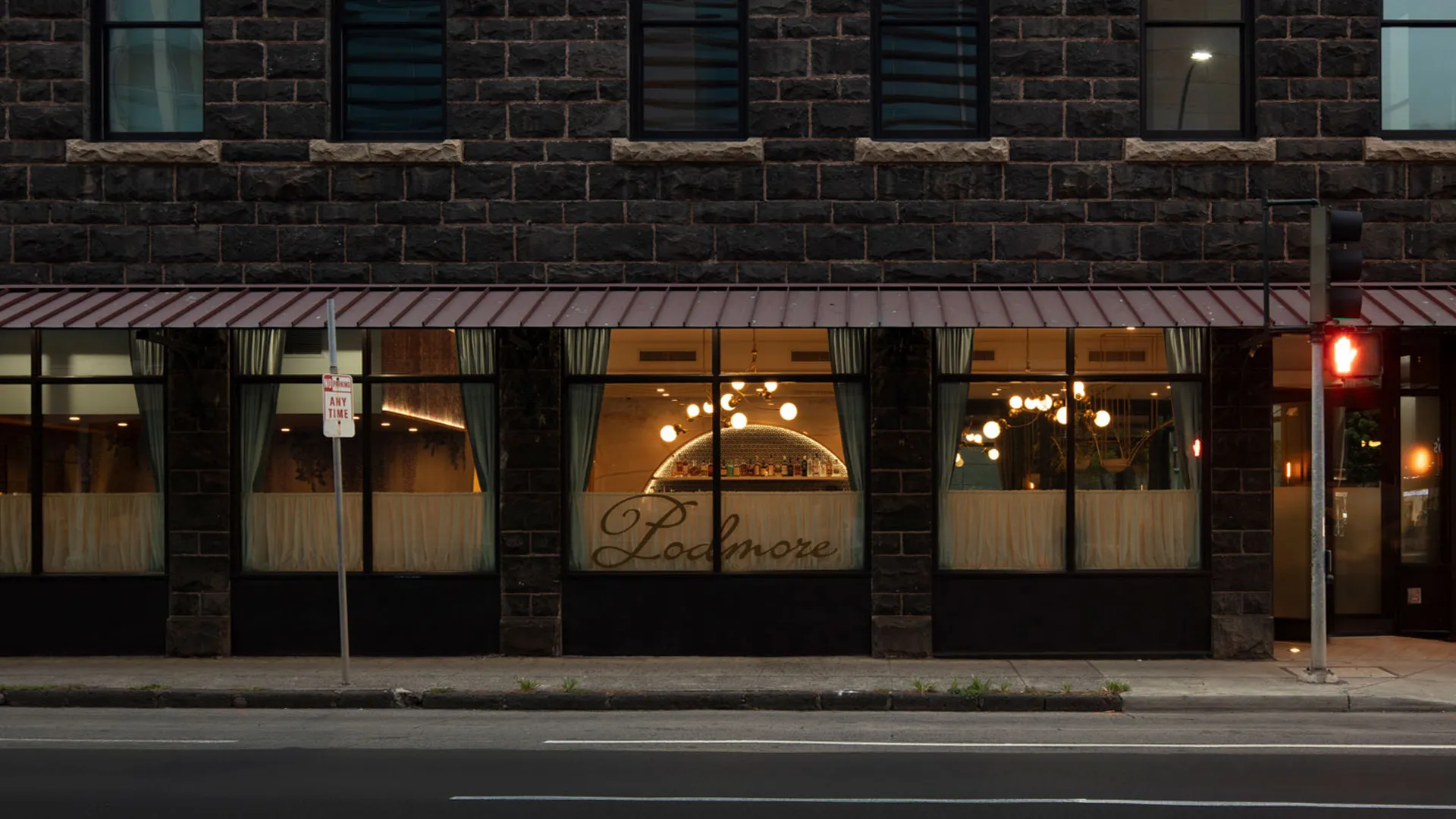 Exterior of Podmore restaurant with lit bar, large windows, and dark brick facade at night.