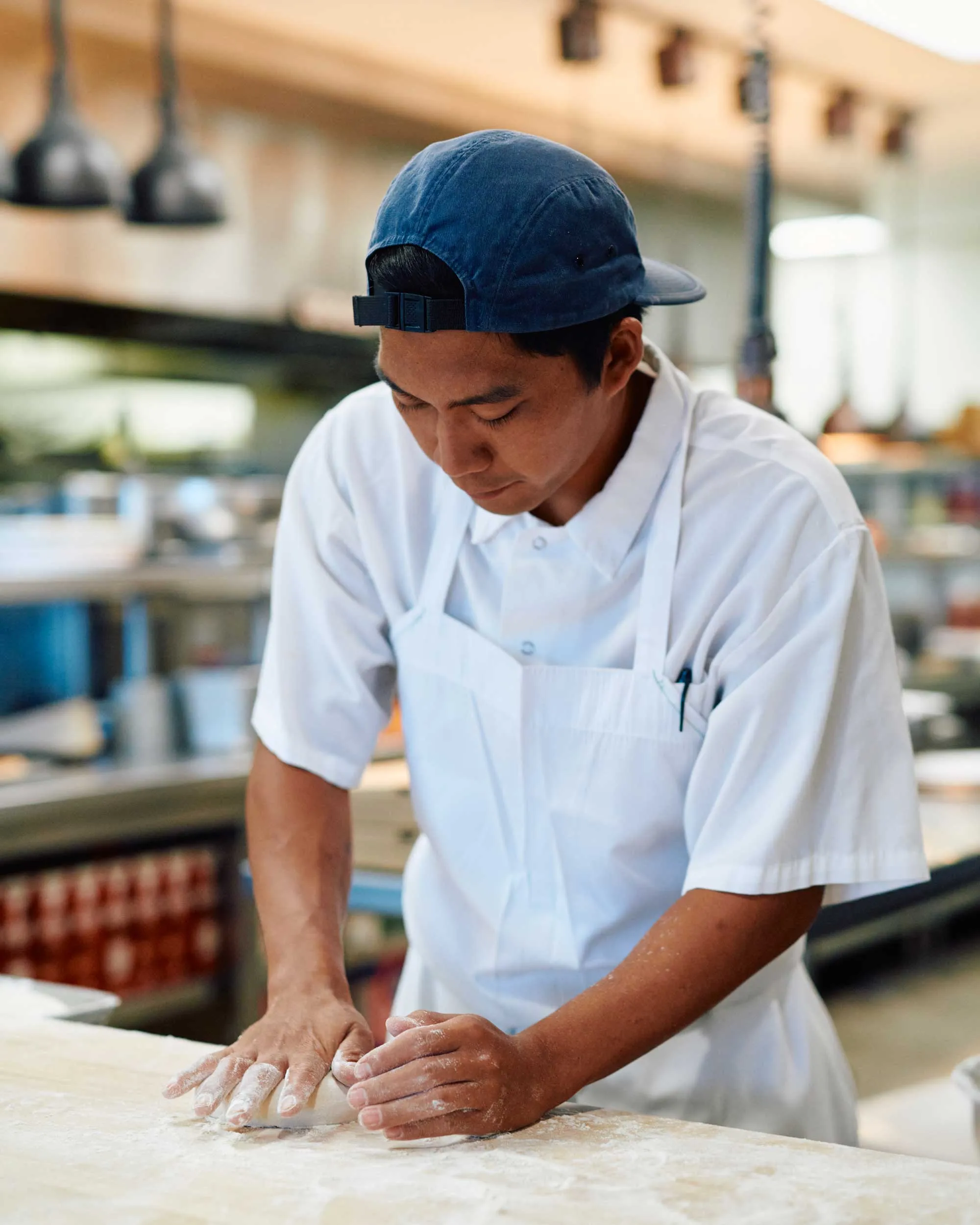 Chef wearing a white apron and blue cap kneading dough on a floured surface in a kitchen.
