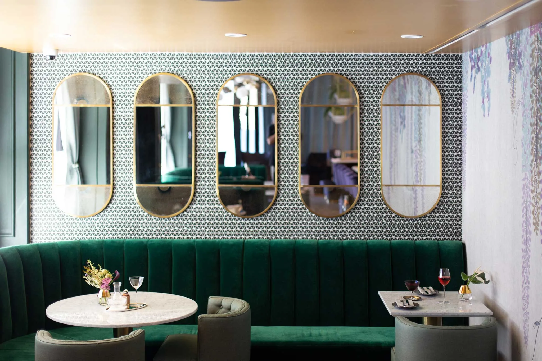 Modern dining area with green velvet bench, marble tables, gray chairs, and five oval mirrors on patterned wall.