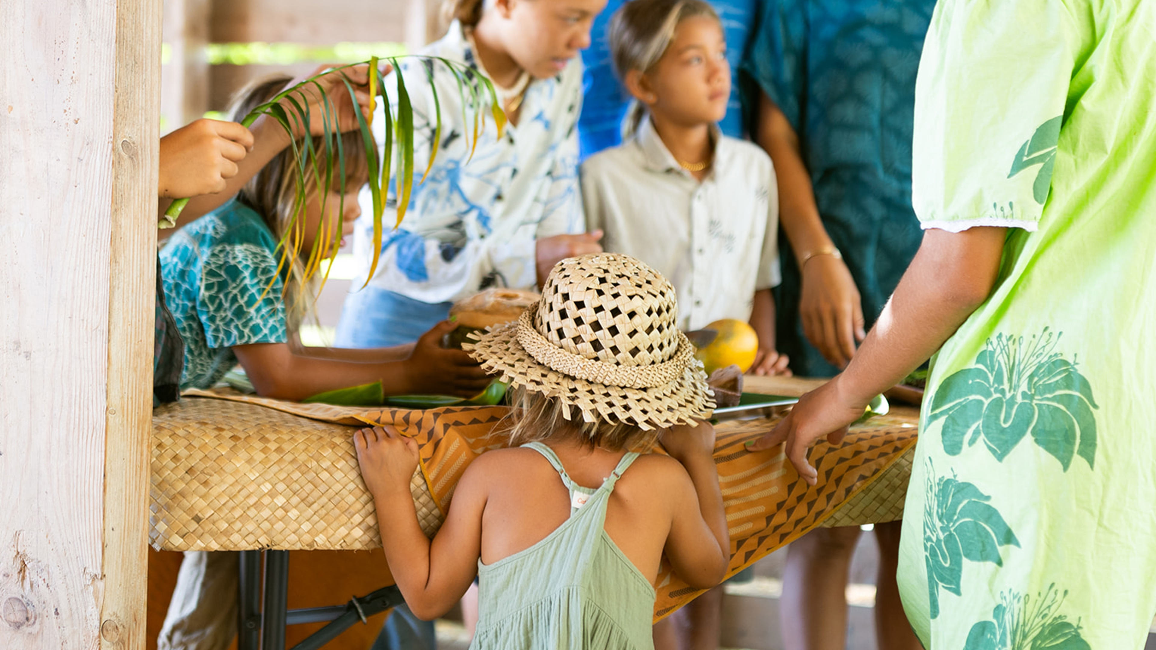 Group of children gathered around a table with fruits and plants indoors, one wearing a straw hat.