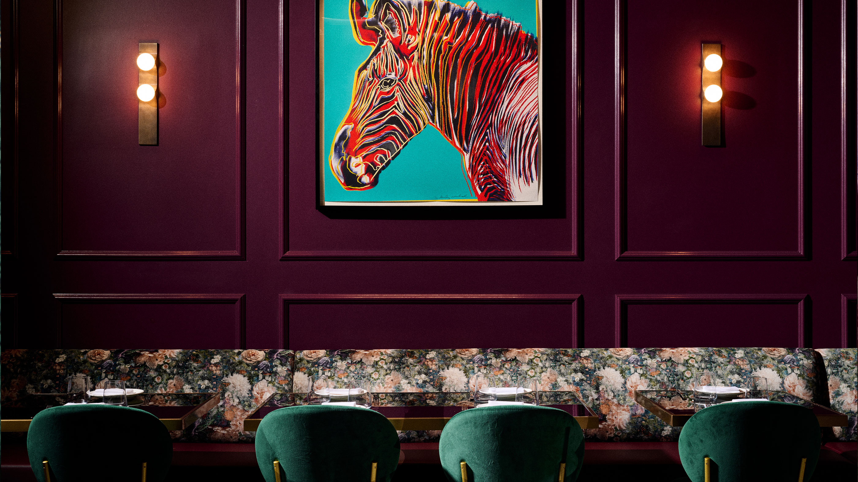 Dining area with floral bench, green chairs, burgundy paneled walls, lit sconces, and colorful zebra artwork.
