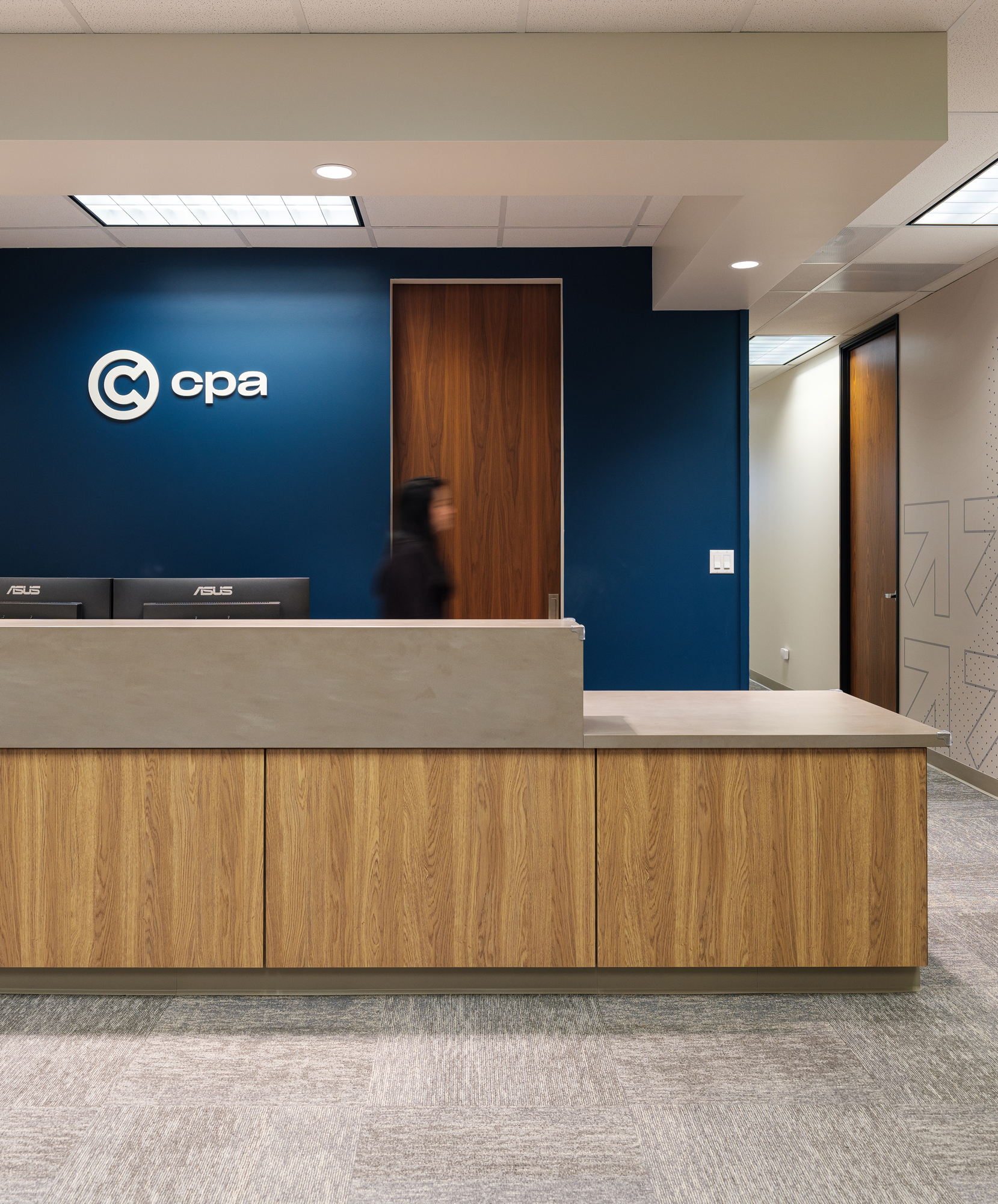 Modern office reception desk with wood panels, blue wall, company logo, and a blurred person behind the desk.