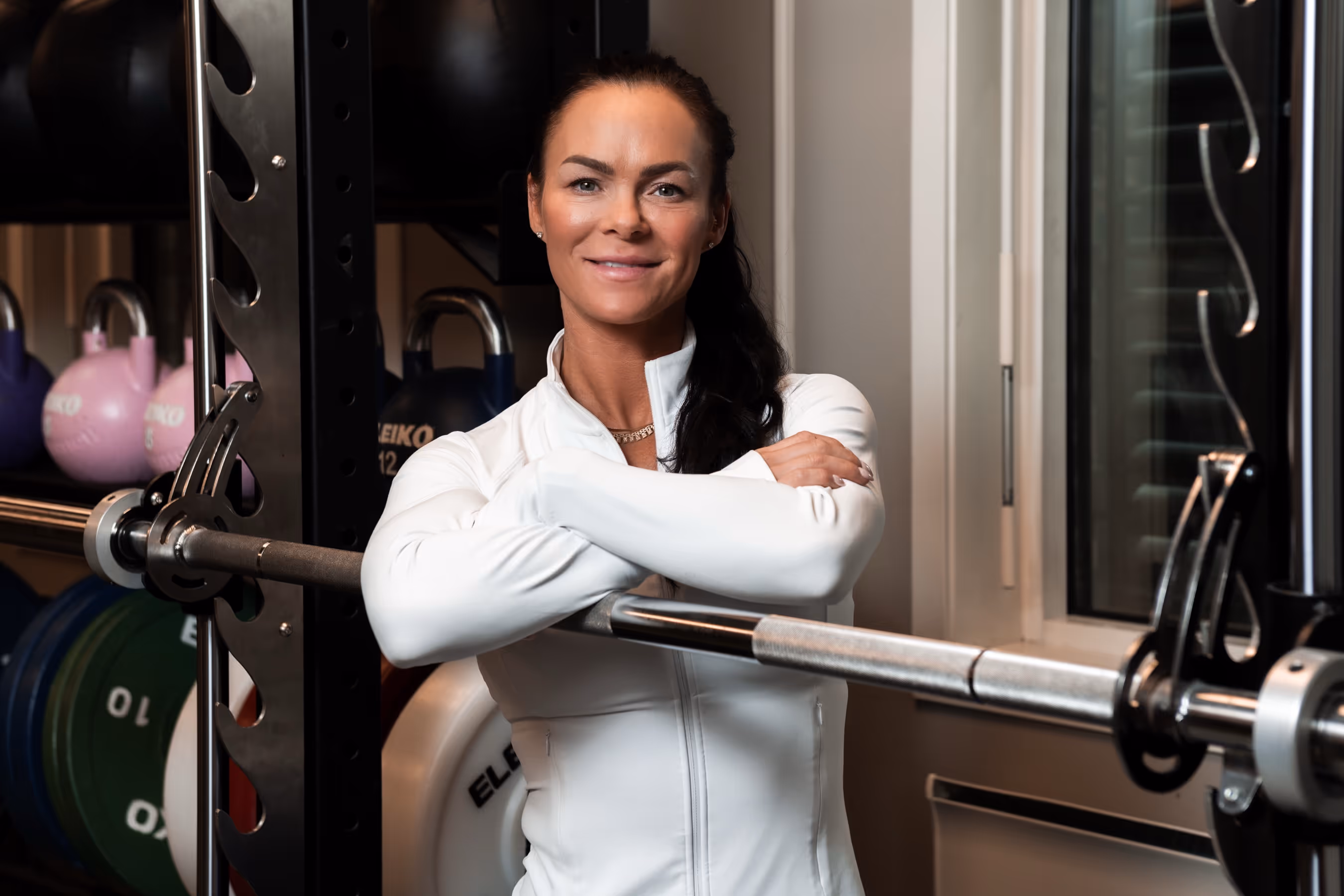 Tonje i hvit treningsjakke smilende med armene i kors foran en vektstang og kettlebells