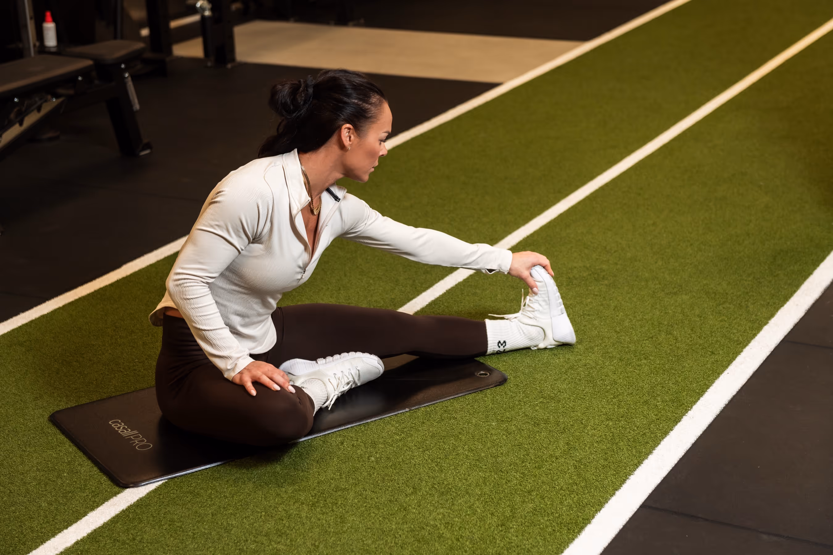 Tonje sitter på en matte og tøyer hamstring mens hun holder foten på kunstgressbane i treningsstudio
