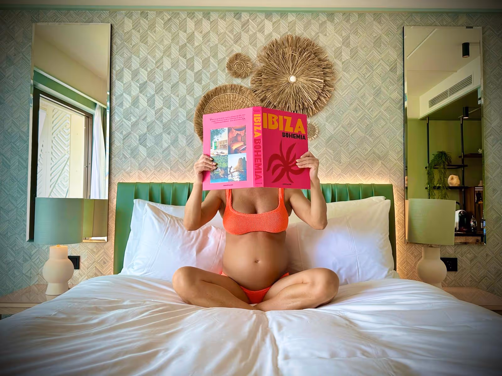 Gravid kvinne i bikini som sitter på en hotellseng og leser boken Ibiza Bohemia foran ansiktet