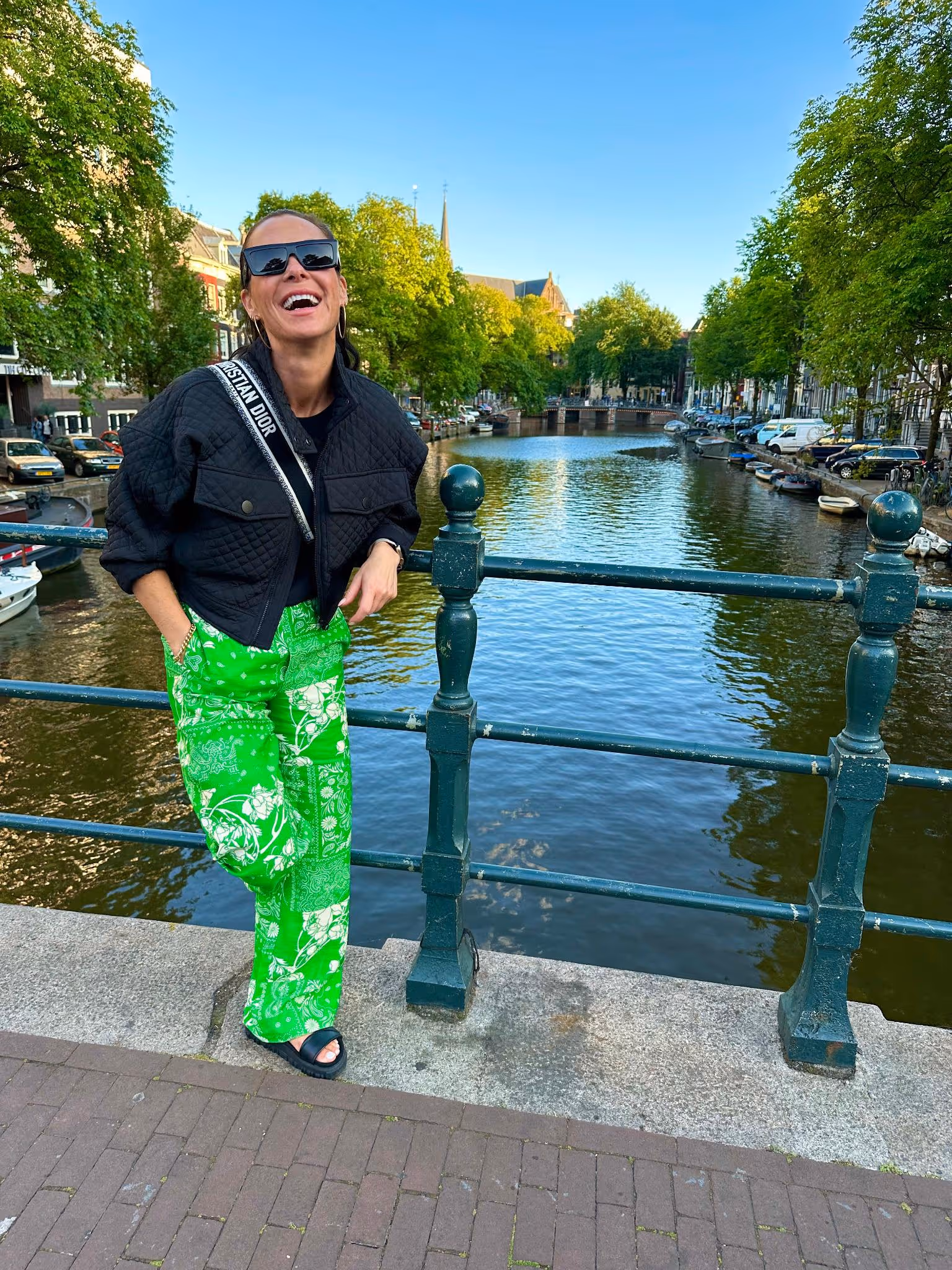Tonje smilende ved en kanalbro i Amsterdam iført solbriller og grønne bukser