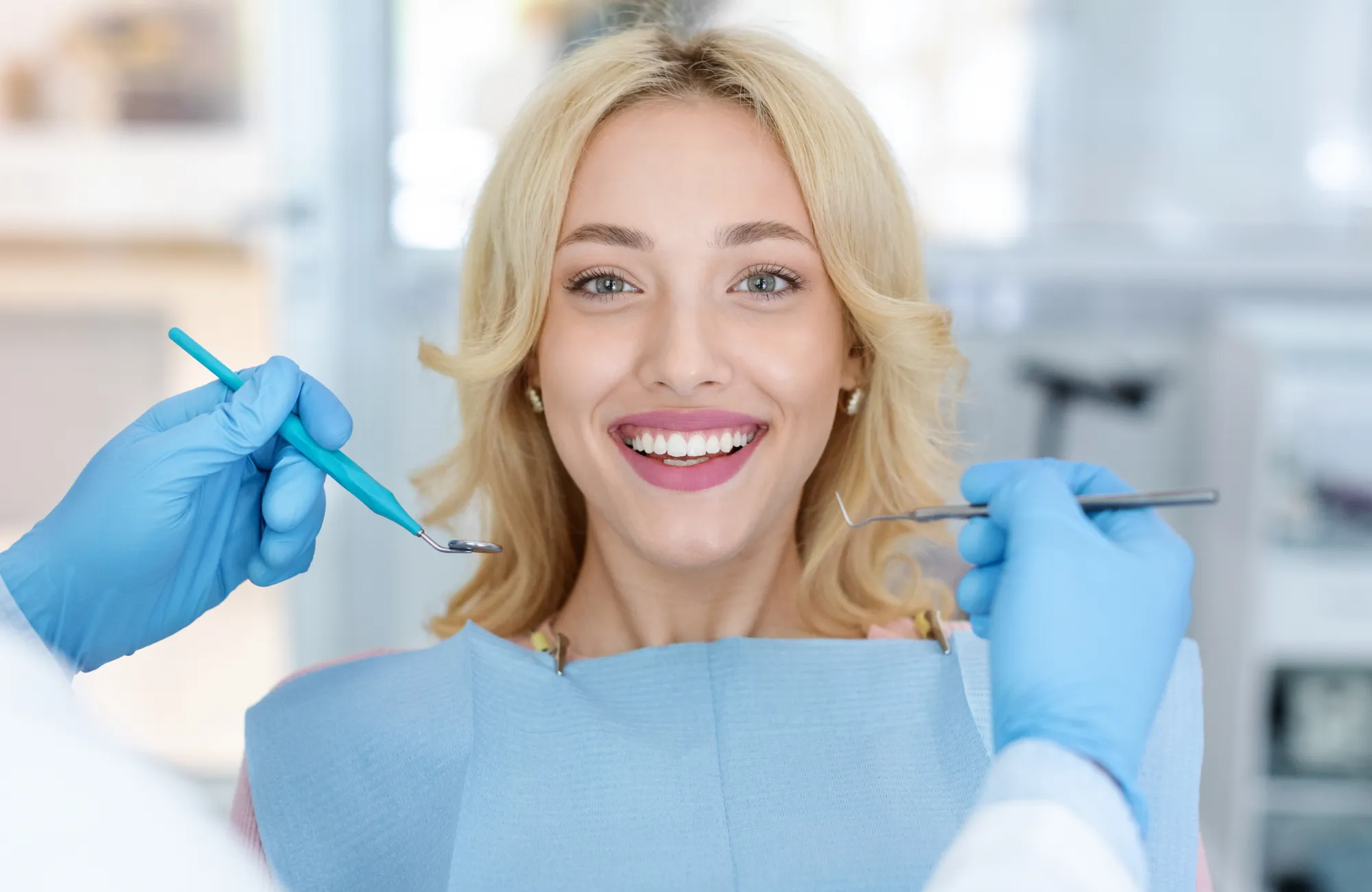 A woman smiles while sitting in a dental chair, surrounded by a dentist's hands holding tools.