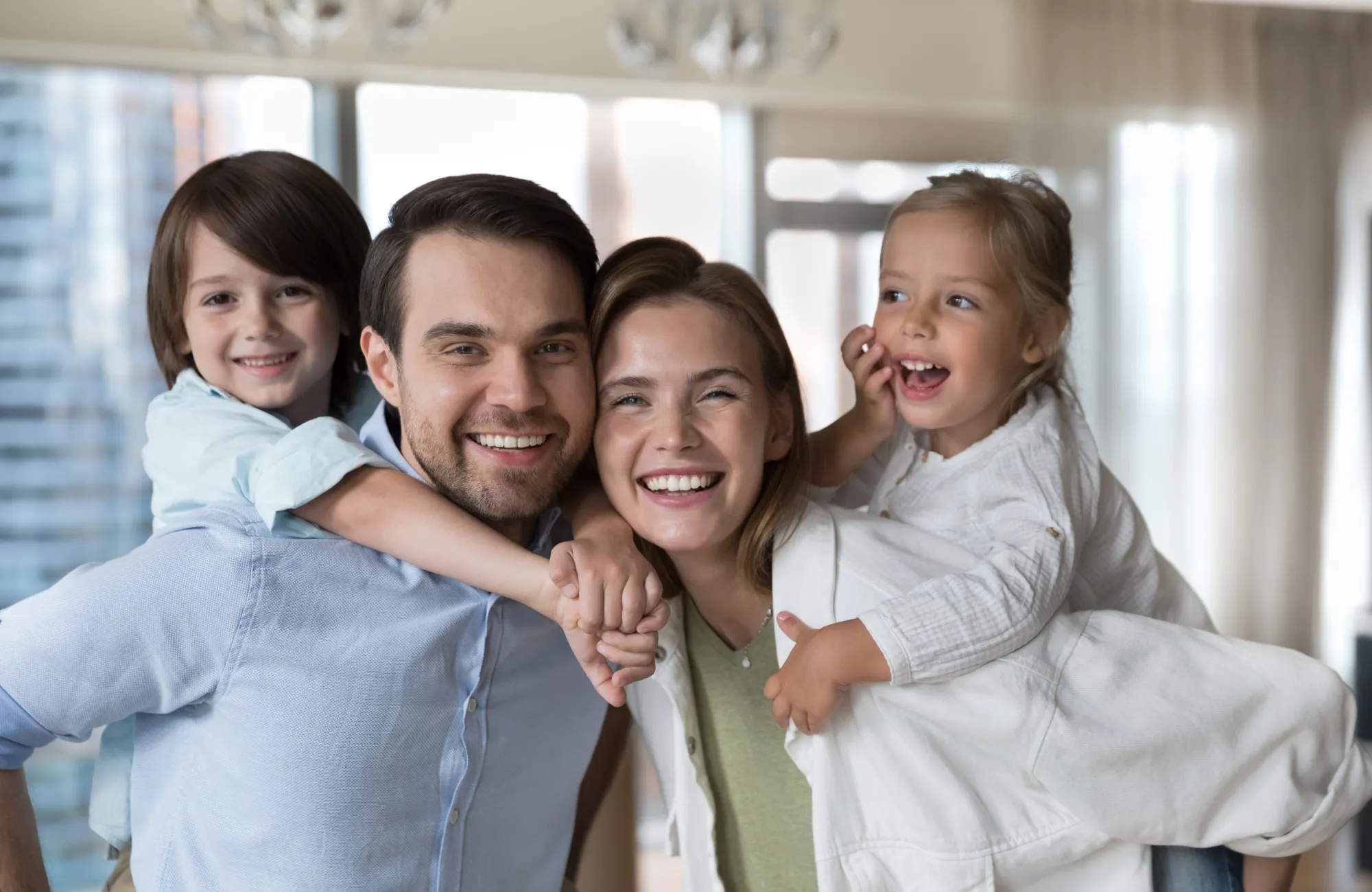 A happy family of four smiling, with parents holding two young children indoors.