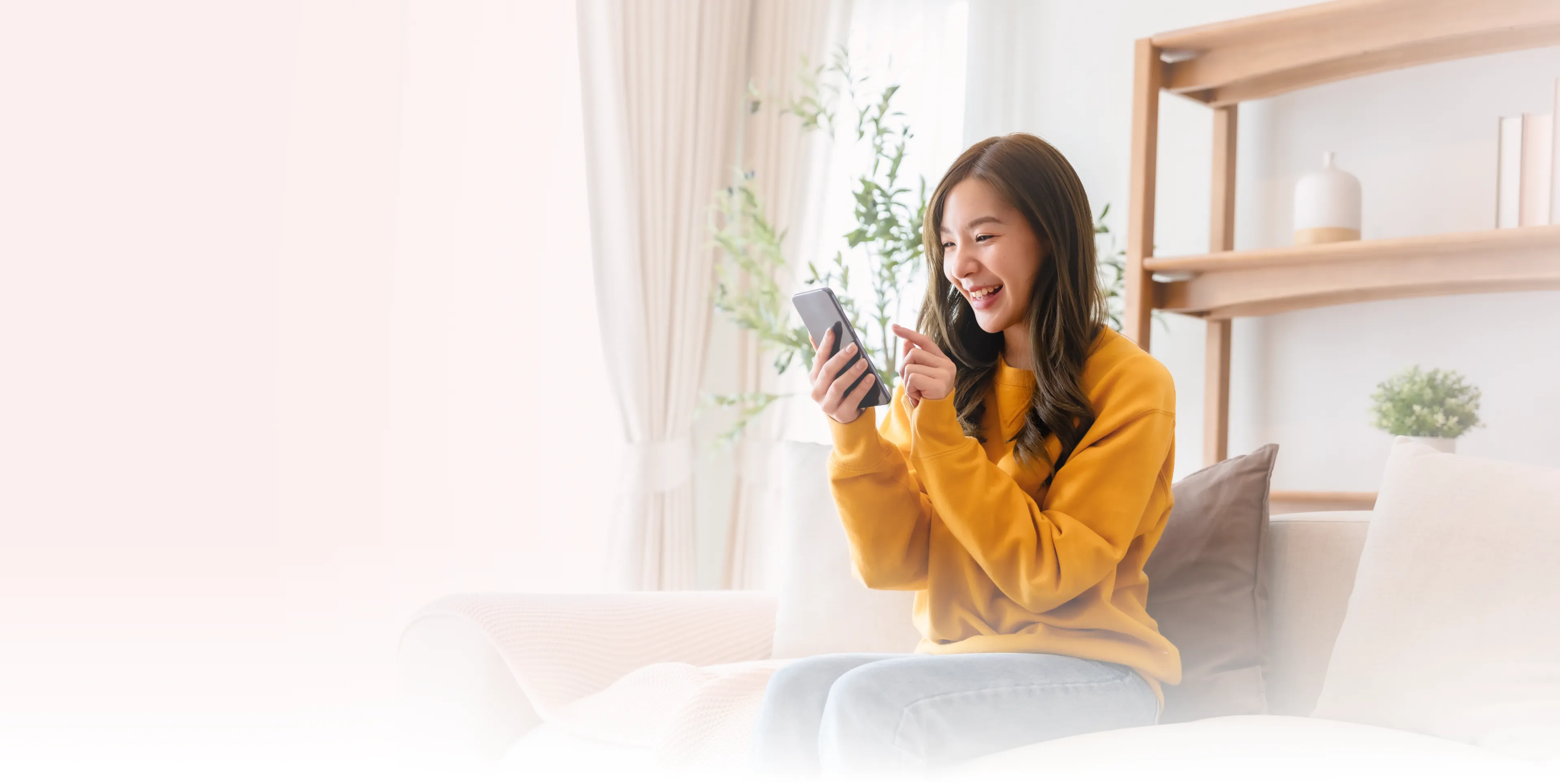 A woman in a yellow sweater sits on a couch, smiling and using her phone.