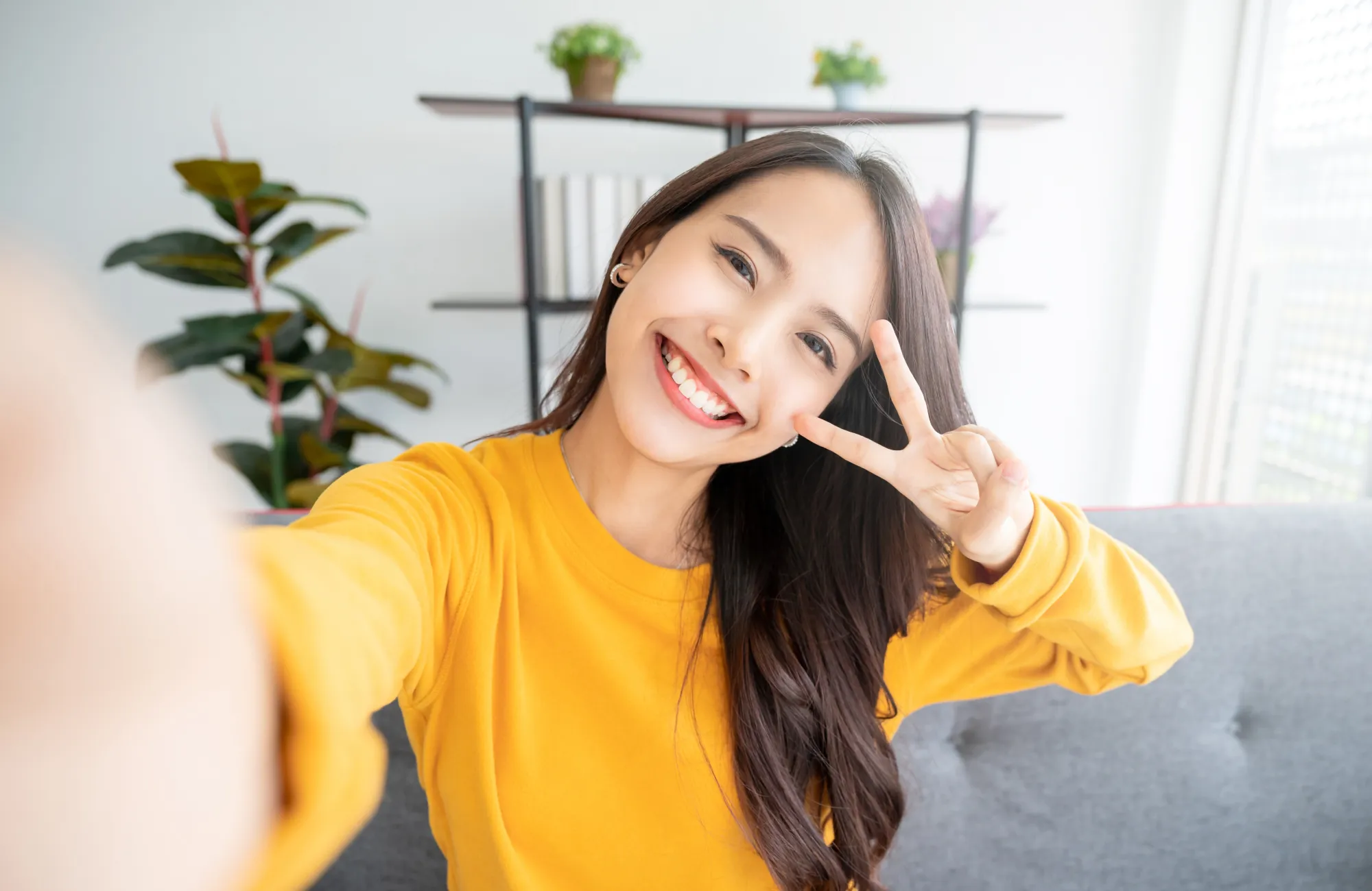 Woman in a yellow sweater smiles and makes a peace sign while taking a selfie indoors.