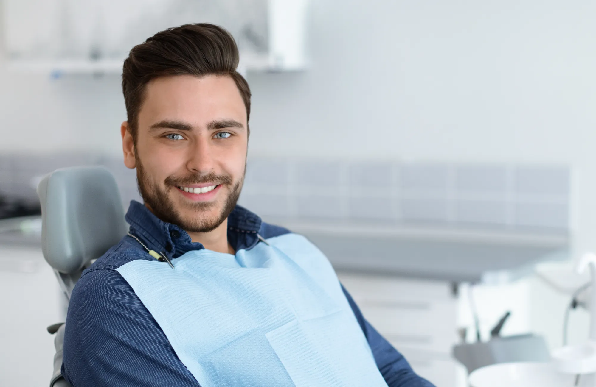 A man sitting in a dentist's chair, wearing a blue dental bib, smiles at the camera.