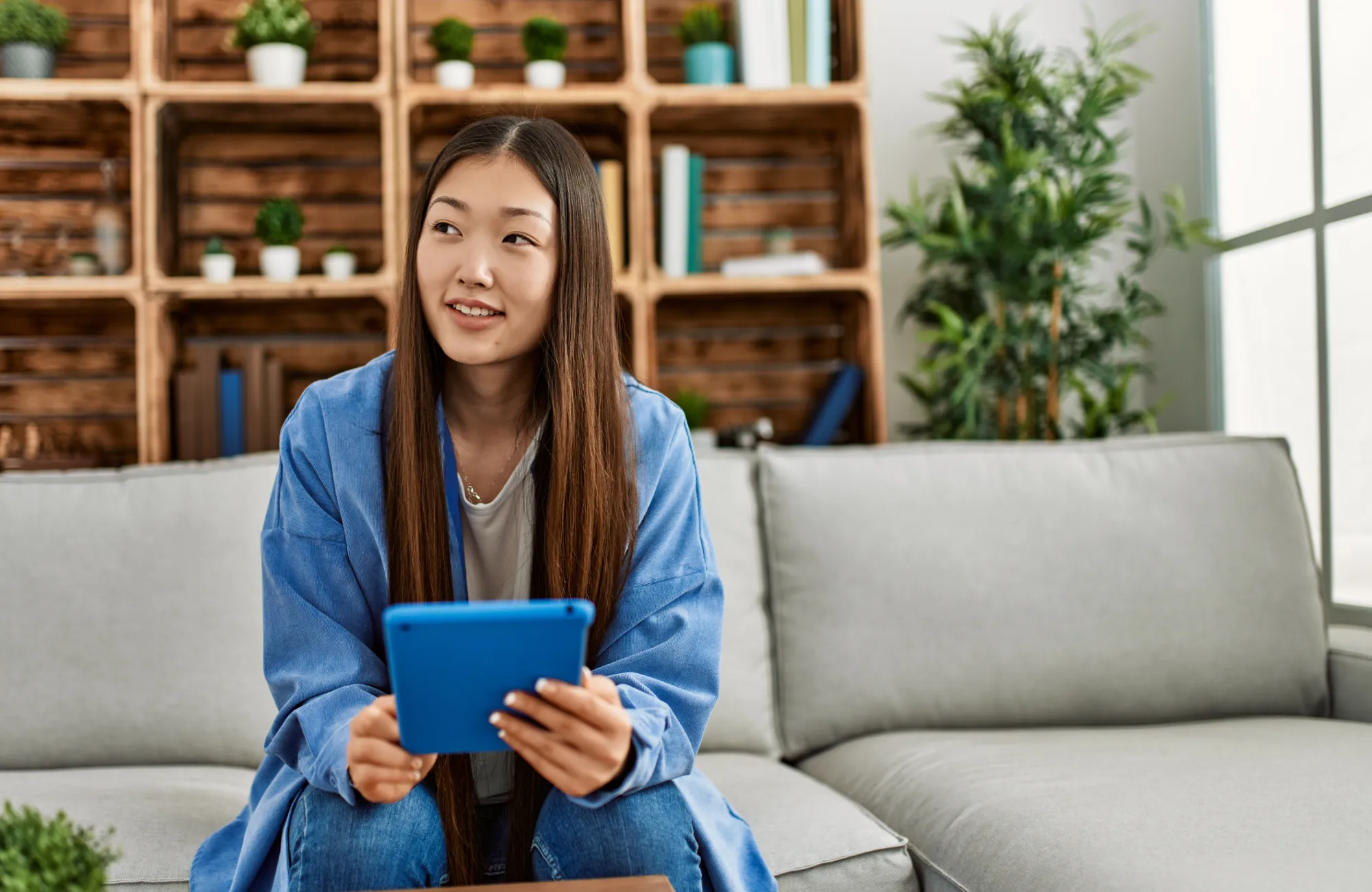 Young woman sitting on a couch, holding a blue tablet and smiling.
