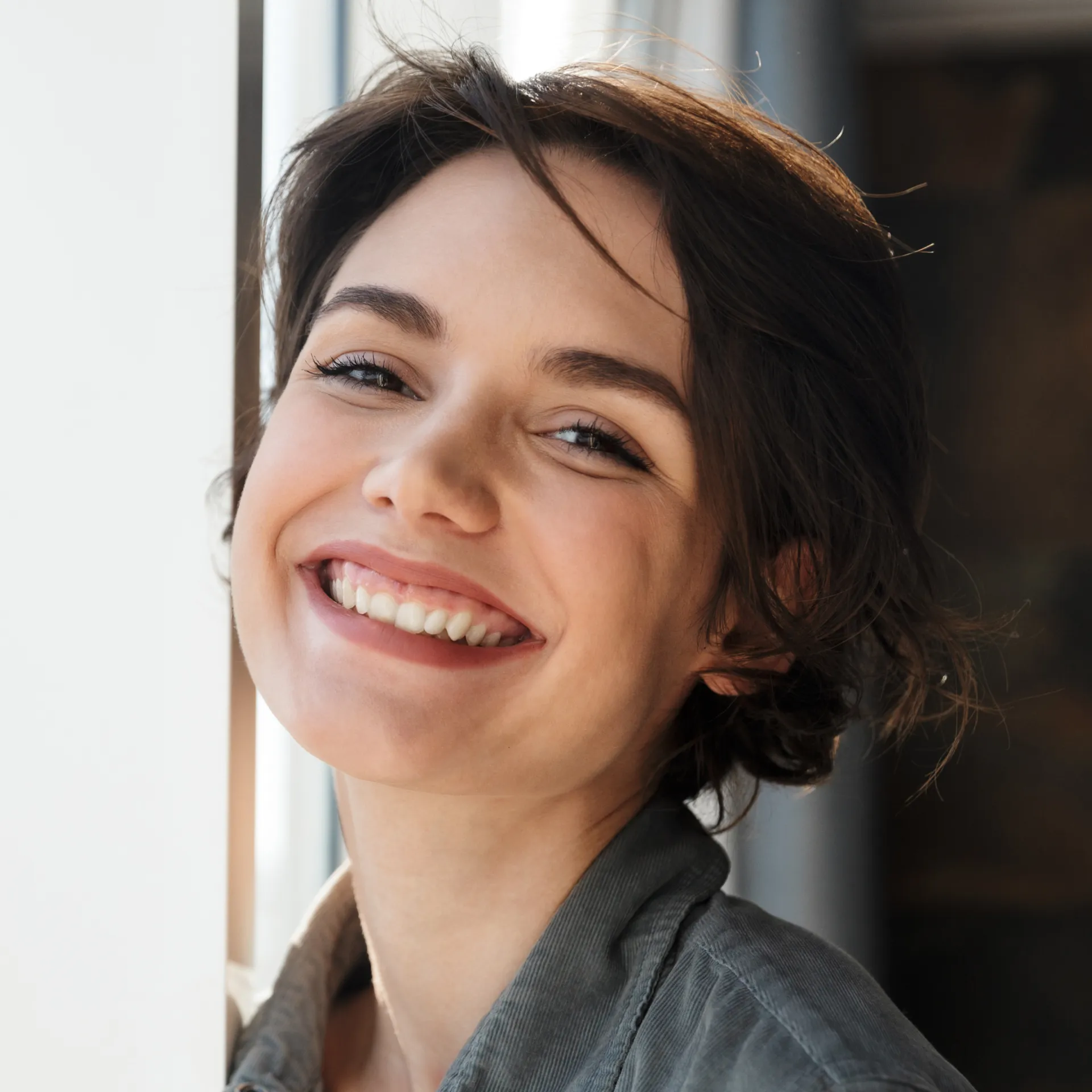 Close-up of a young woman with short brown hair smiling warmly by a window.