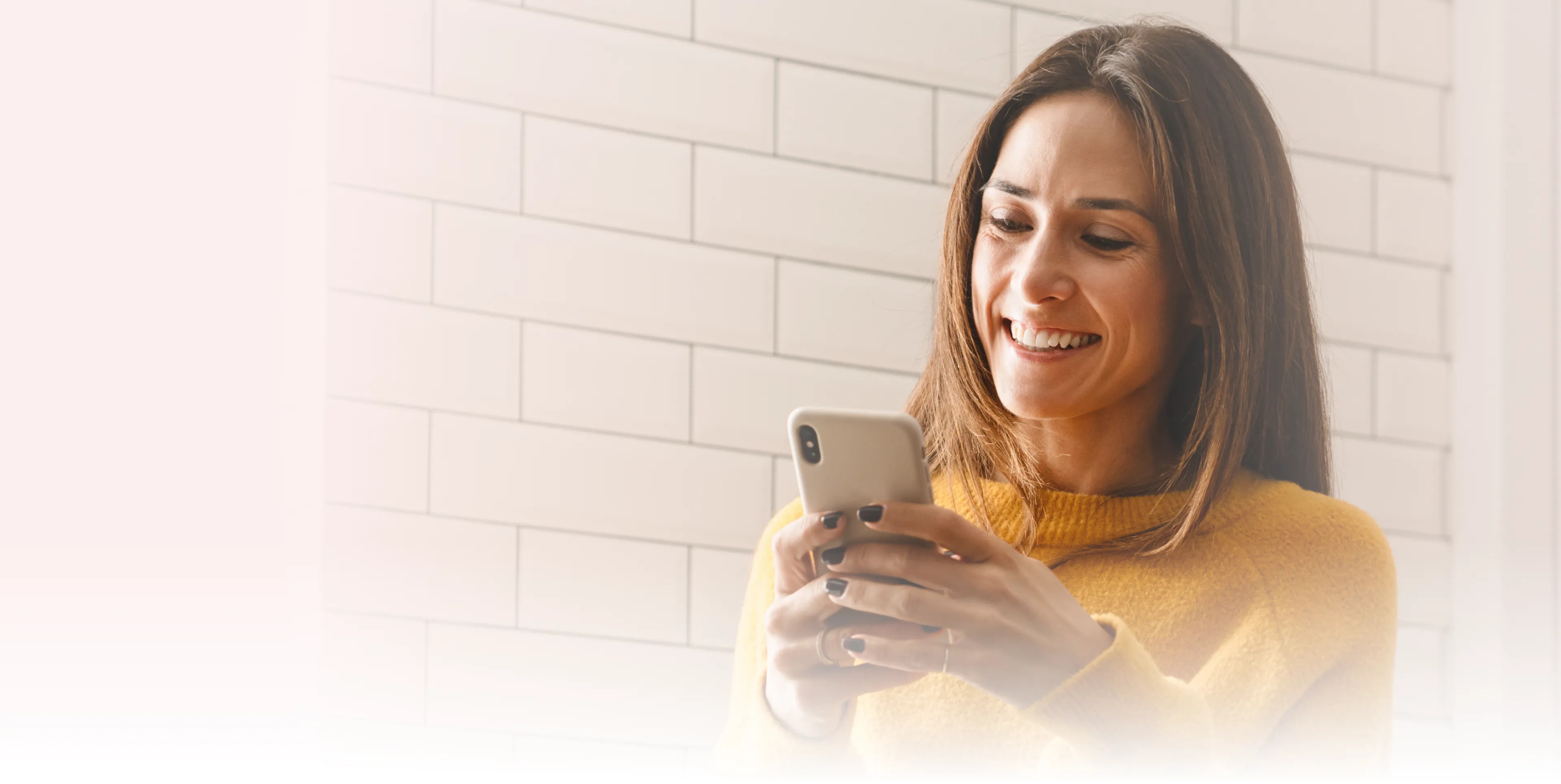 A woman in a yellow sweater smiles while looking at her smartphone.