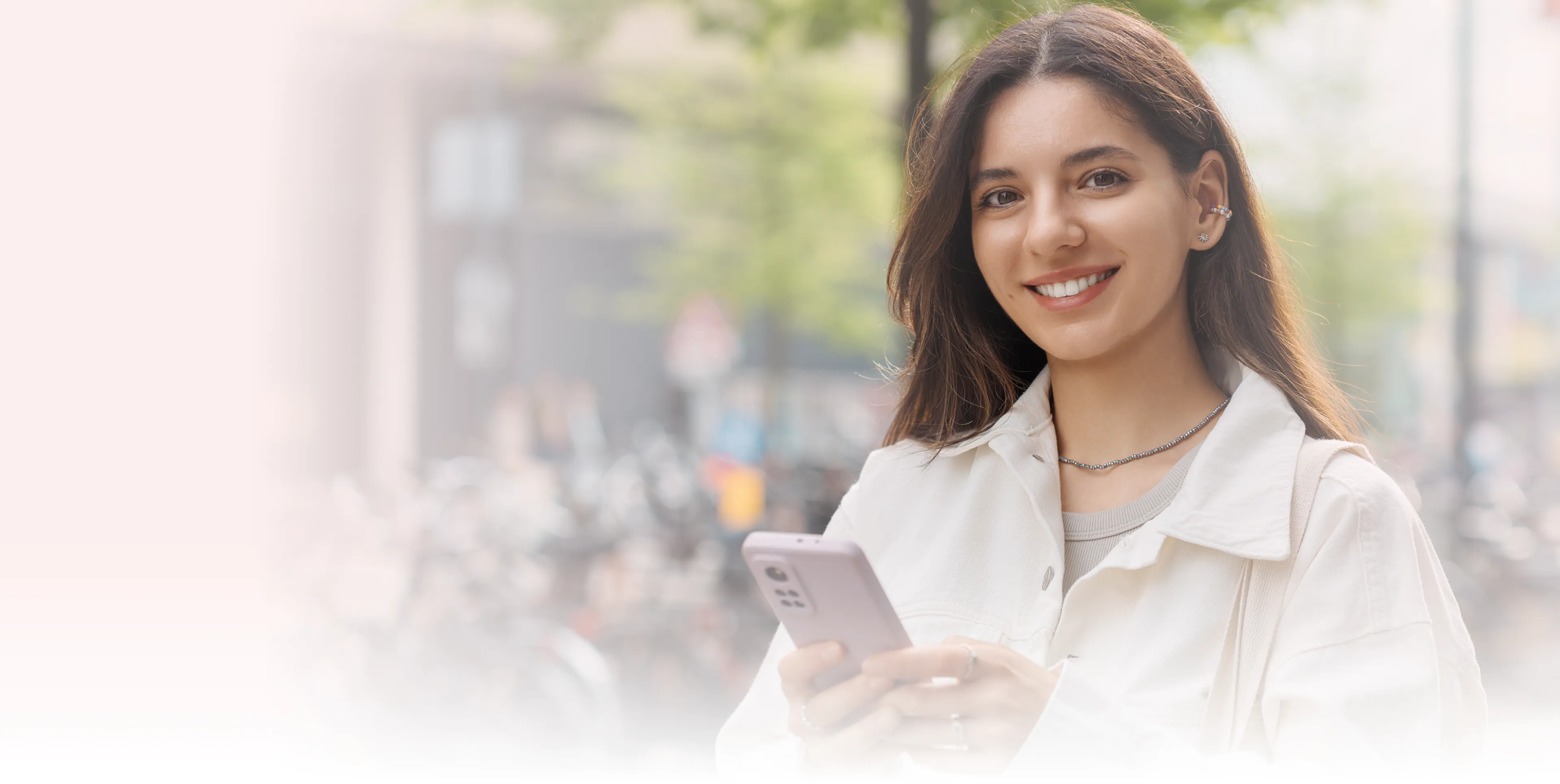 A woman smiles while holding a smartphone outdoors.