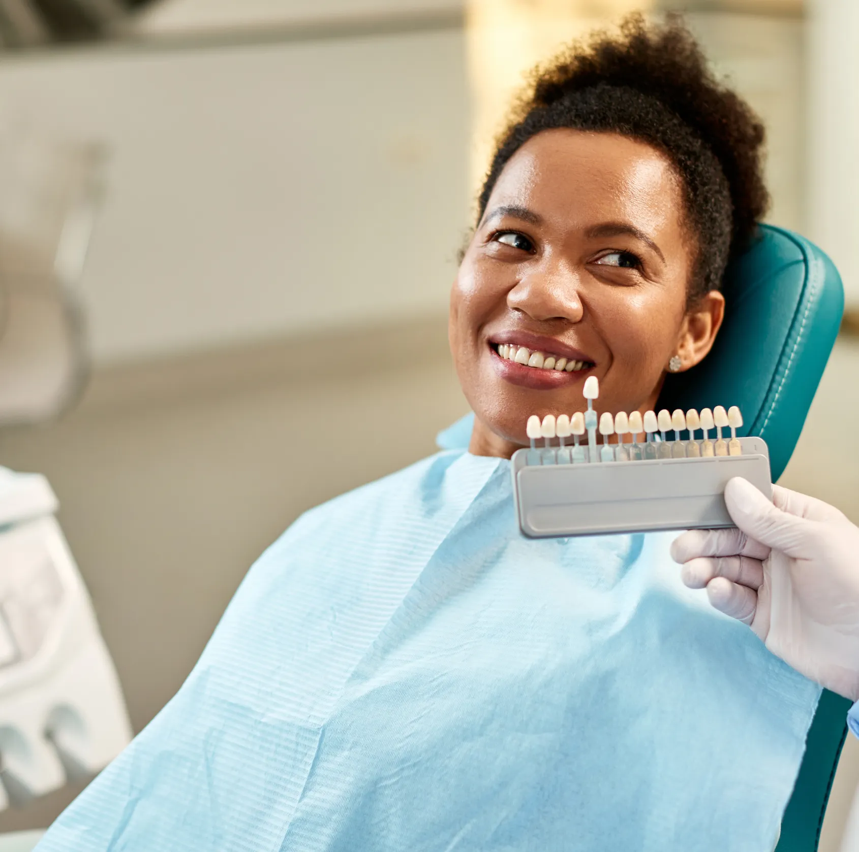 A person in a dental chair smiles while a dentist shows a tooth color shade guide.