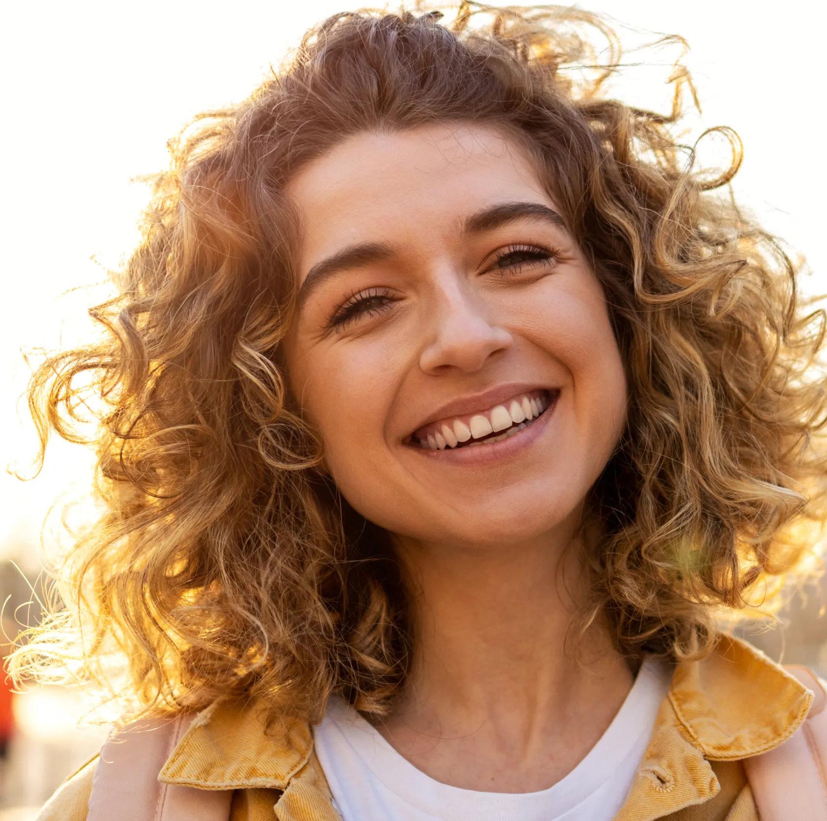 A person with curly hair smiles warmly at the camera in sunlight.