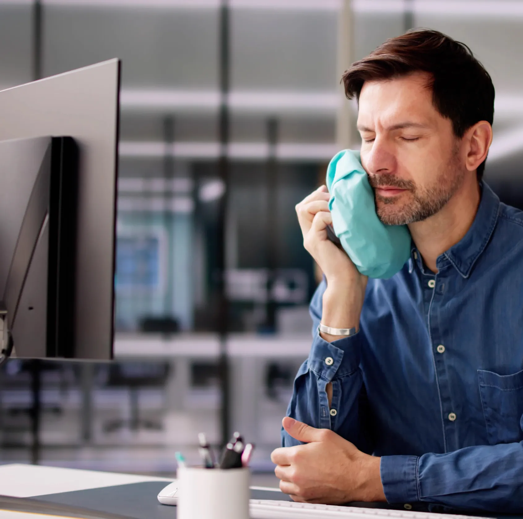 A man sitting at a desk applies an ice pack to his face while looking at a computer.