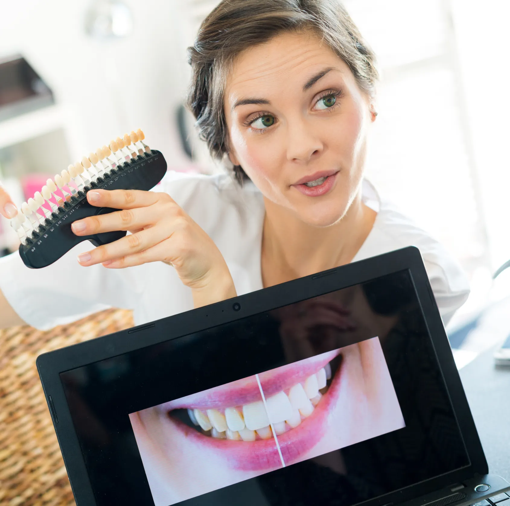 A dental professional holds a tooth color chart, showing a dental image on a laptop screen.