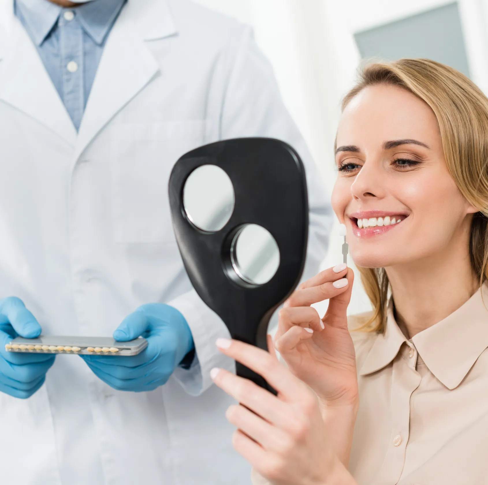 A woman smiles in a dental office, holding a tooth shade guide in front of a mirror.