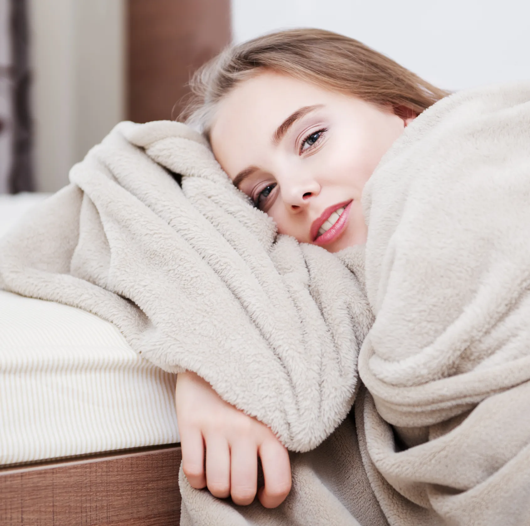 A person wrapped in a blanket, lying on a bed, and smiling at the camera.