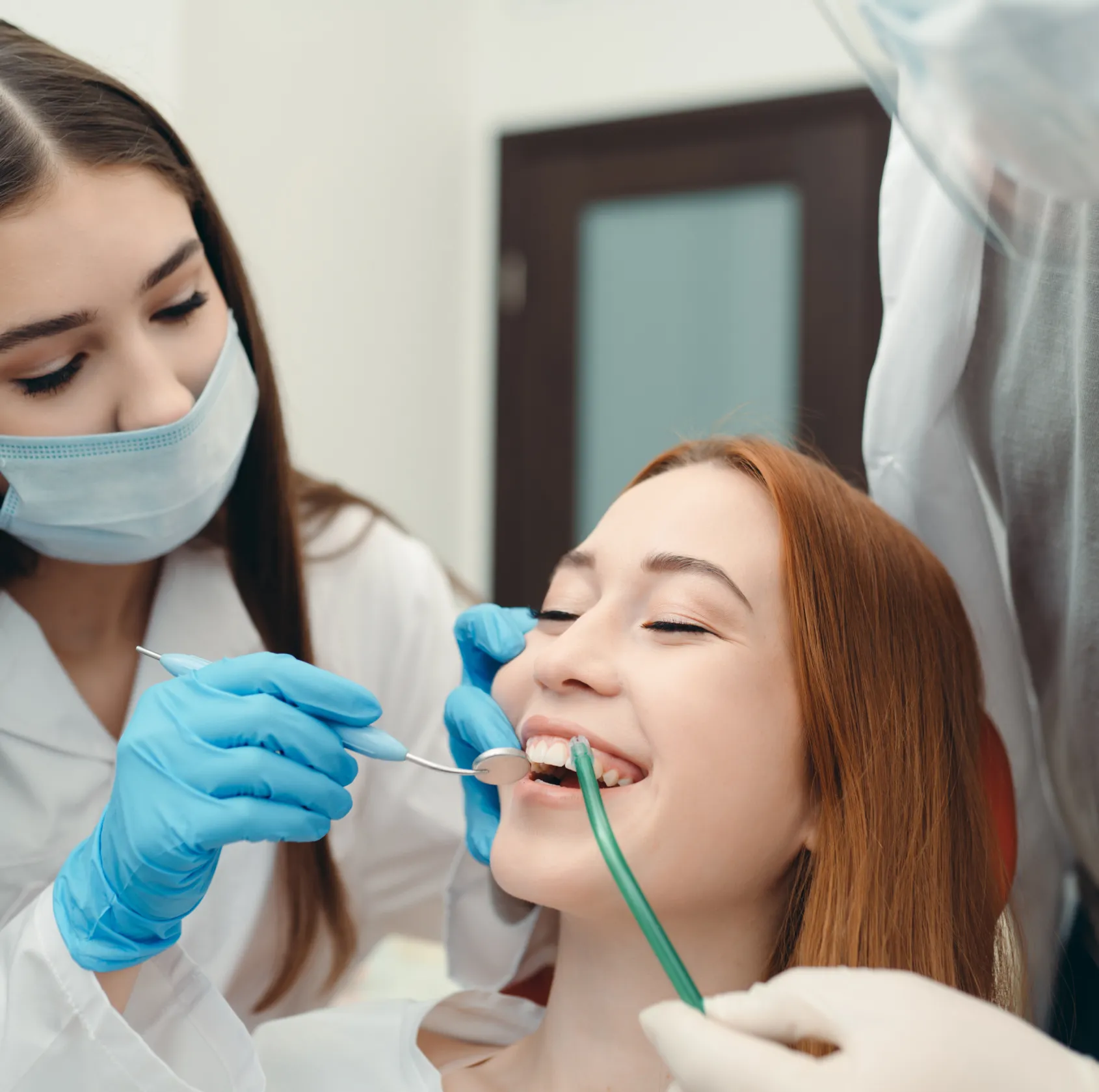 A woman sits in a dental chair, smiling, while a dentist examines her teeth with tools.