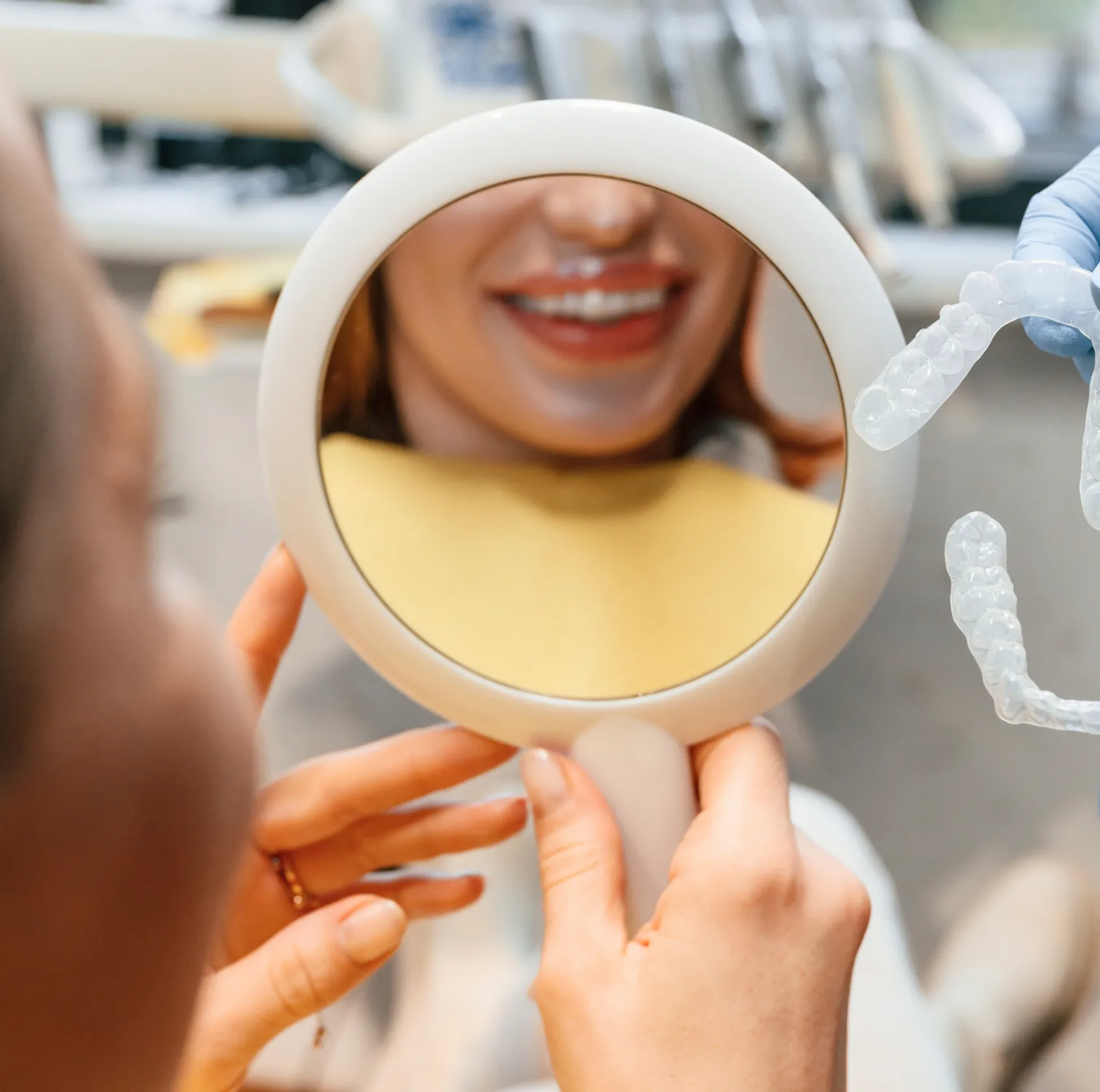 A person smiles while holding a mirror, with a dentist showing them a dental model.