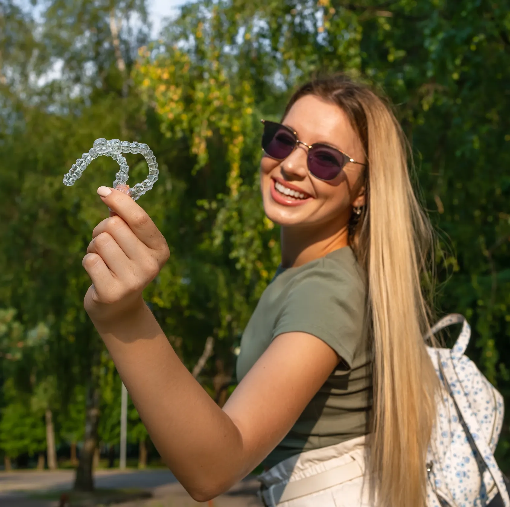 Woman smiling and holding clear dental braces outdoors, wearing sunglasses and a casual outfit.