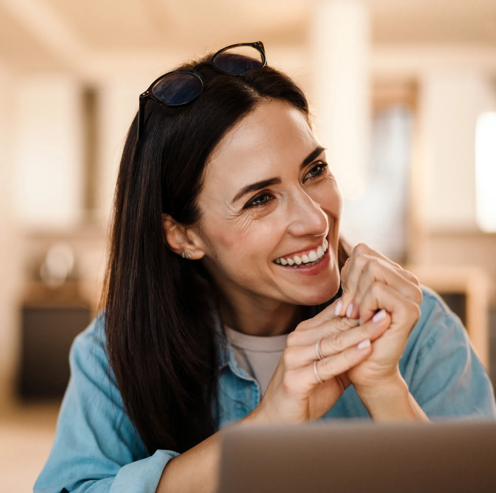 A woman with long dark hair smiles while sitting at a table, wearing glasses on her head.