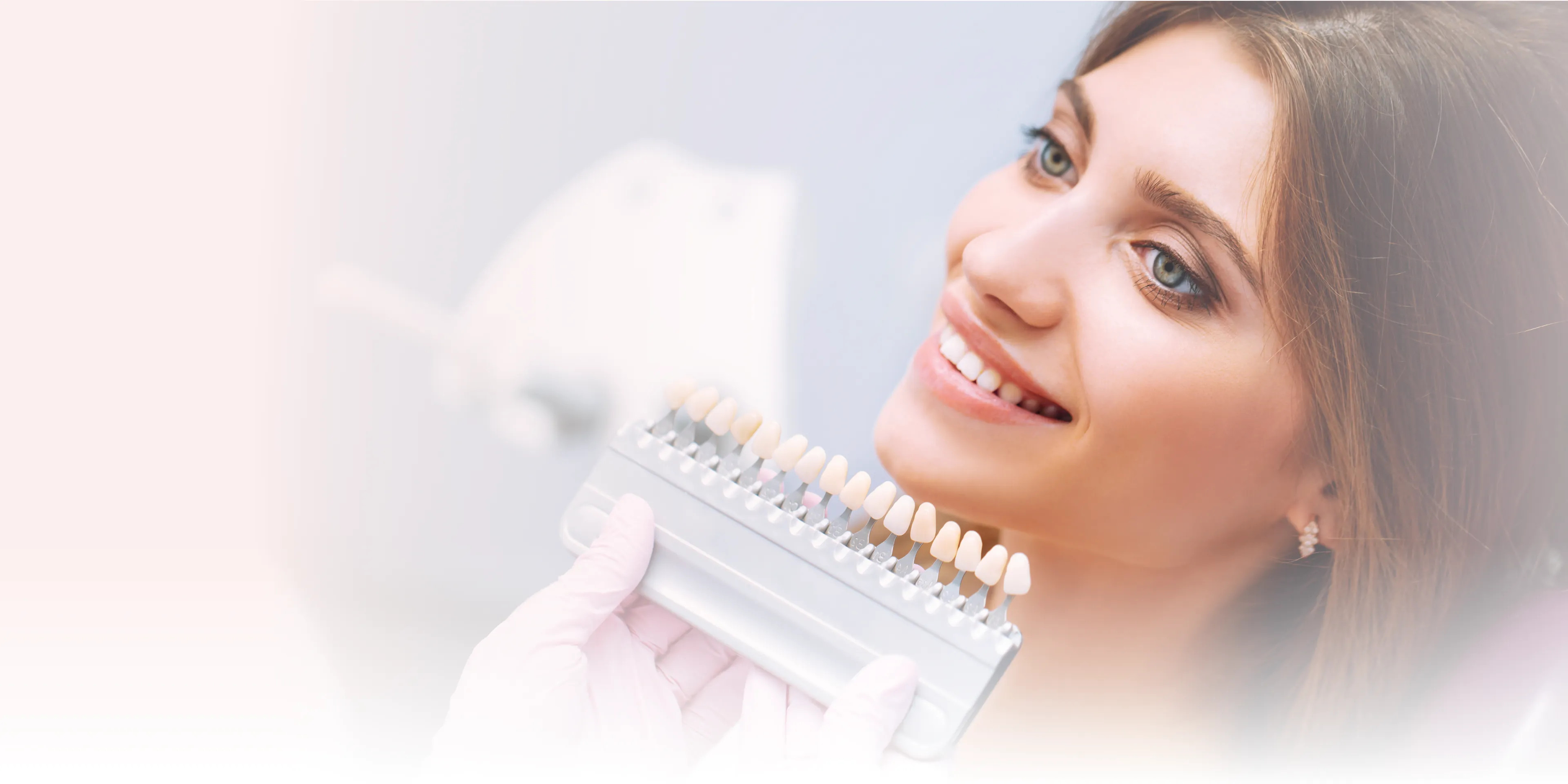 A woman smiles while a dentist holds a dental shade guide near her teeth.