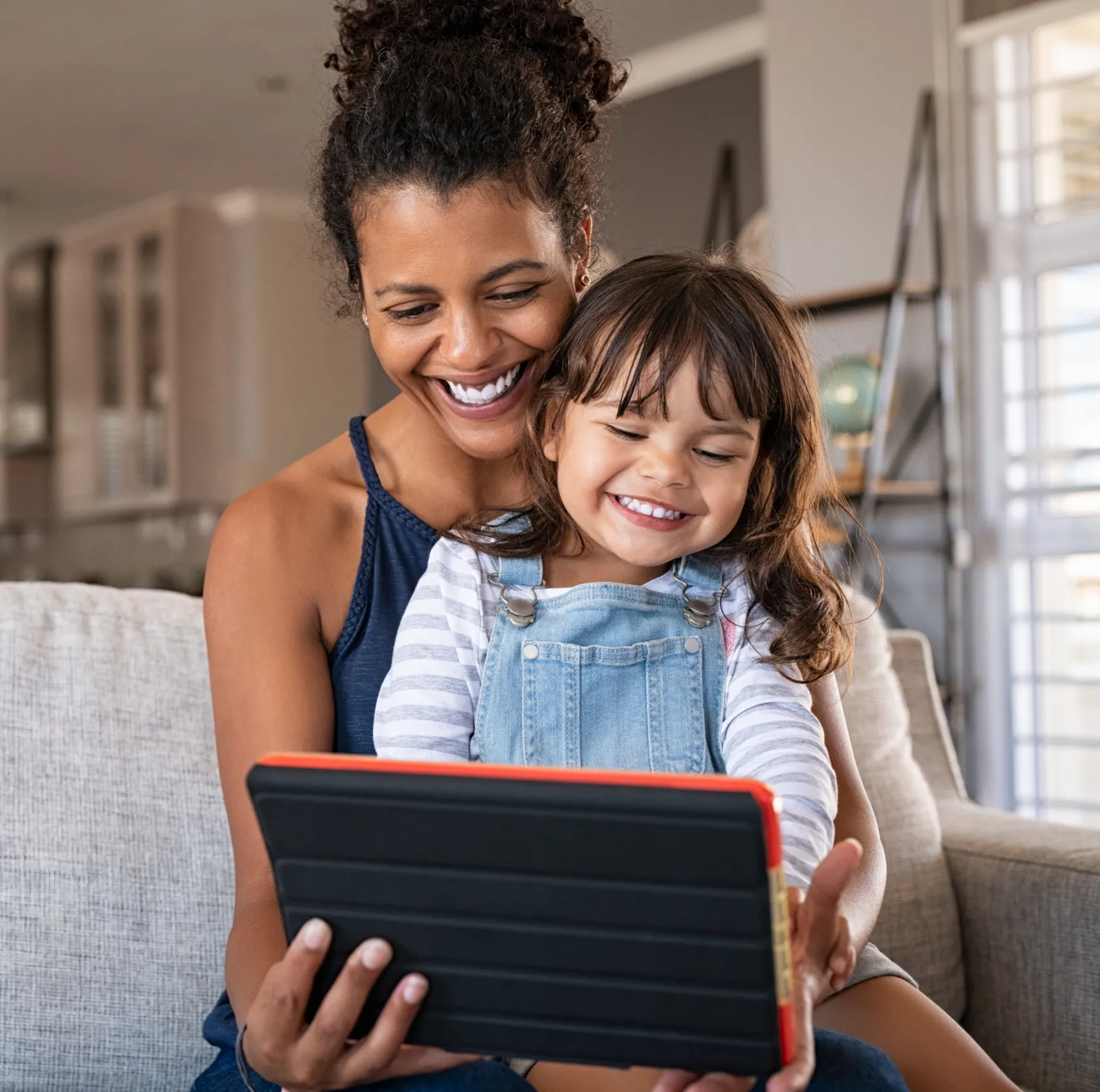 Woman and young girl smiling while looking at a tablet on a couch.