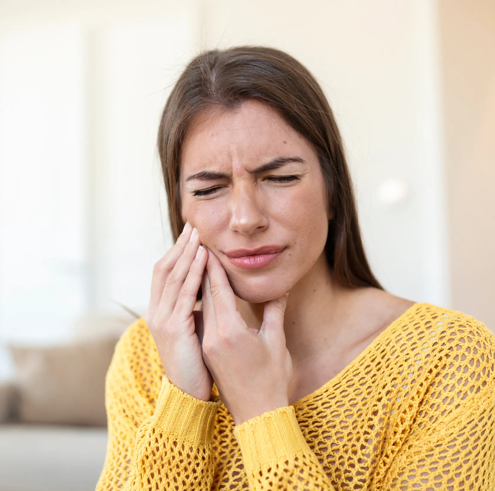 A woman in a yellow sweater touches her cheek, showing signs of tooth pain or discomfort.