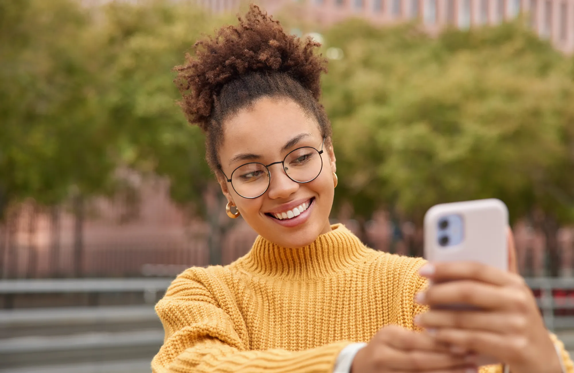 A woman in a yellow sweater takes a selfie with her smartphone, smiling outdoors.