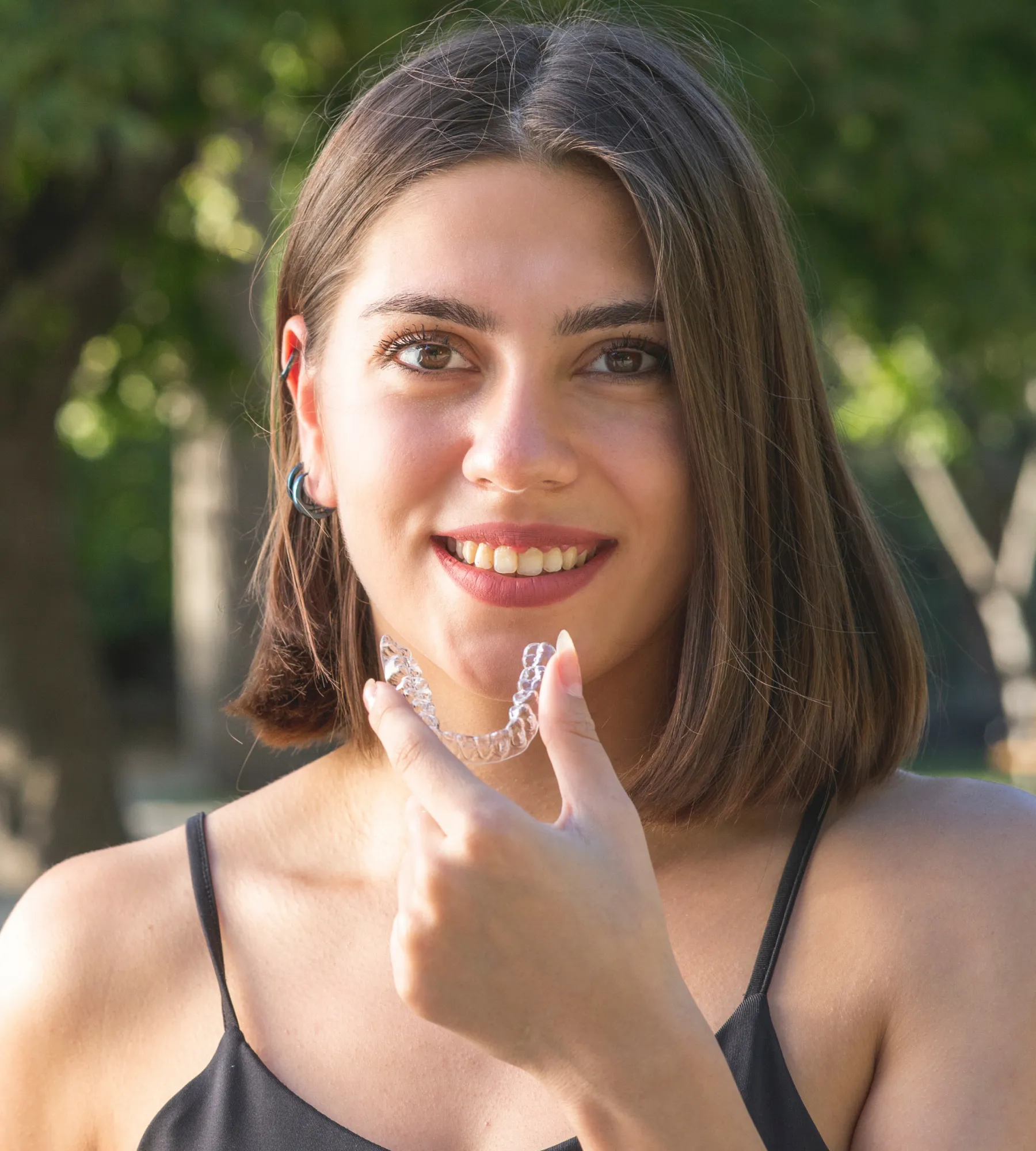 A woman smiles while holding a clear dental retainer near her mouth.