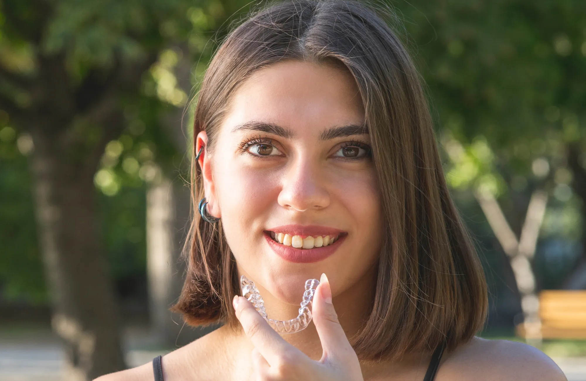 A woman holds a clear dental retainer, smiling outdoors.