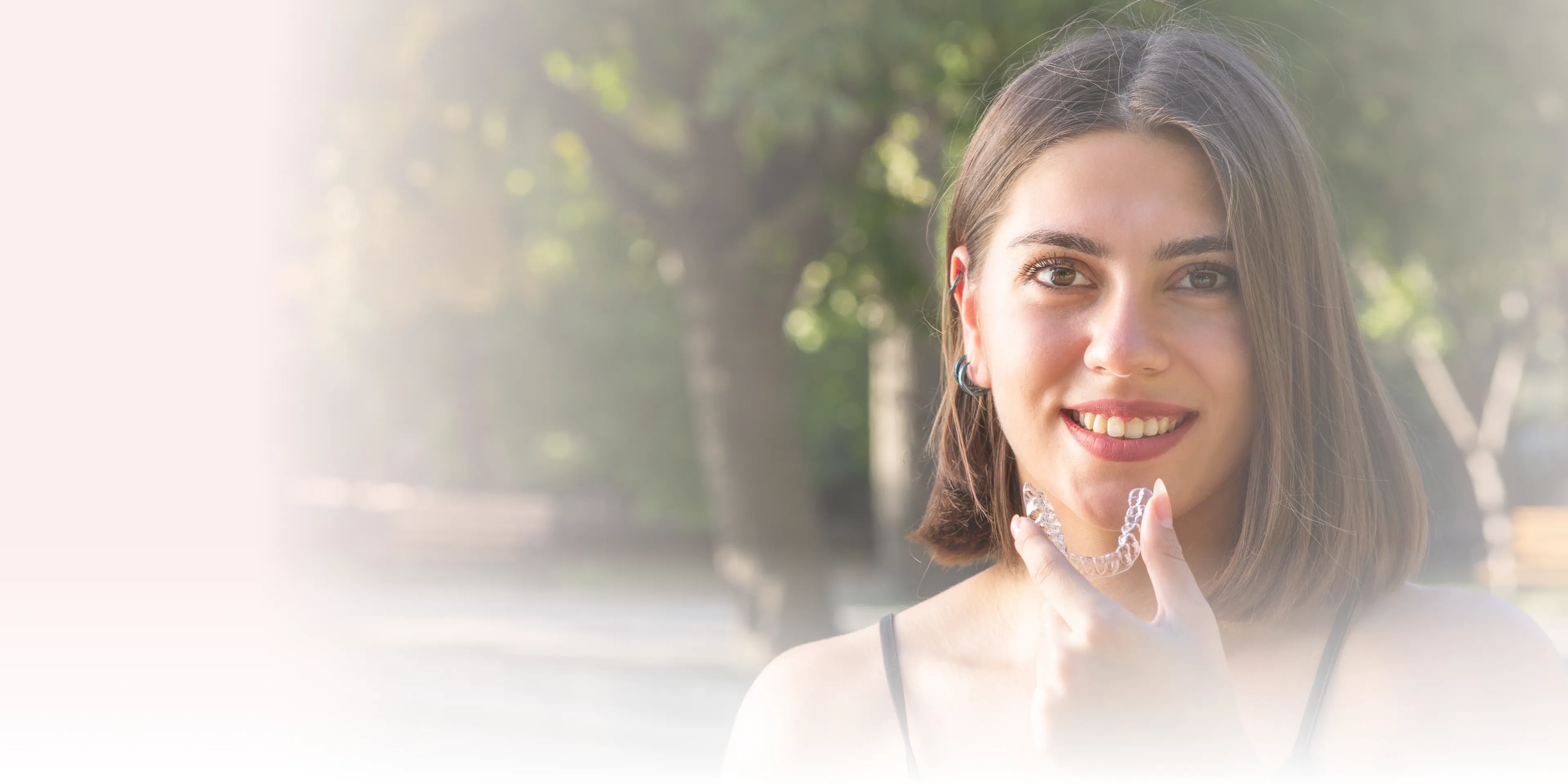 Young woman smiling outdoors holding a clear dental aligner near her mouth.