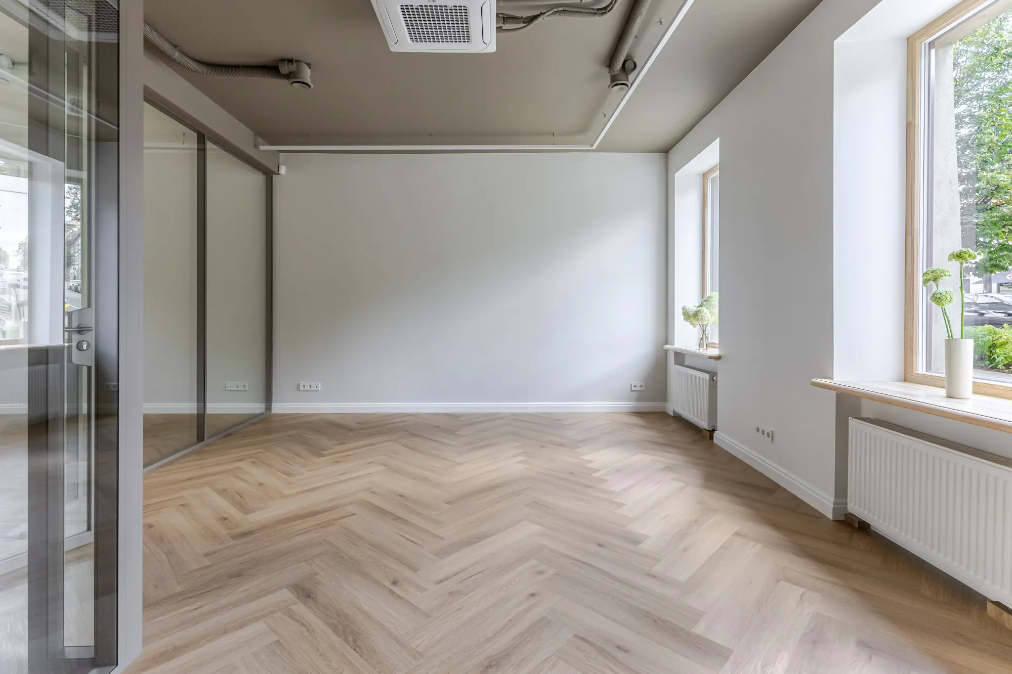 Tuščia modernaus interjero kambario erdvė su šviesiomis medinėmis grindimis, baltais langų rėmais ir vazoniniu augalu ant palangės.