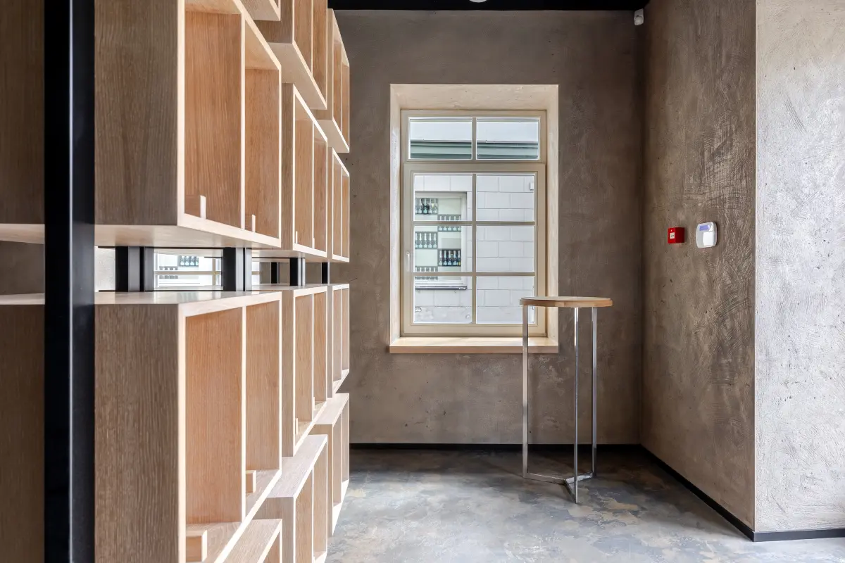 Šiuolaikinio interjero erdvė su medinėmis nišomis palei kairiąją sieną, langas ir aukštas staliukas prie dešinės sienos.