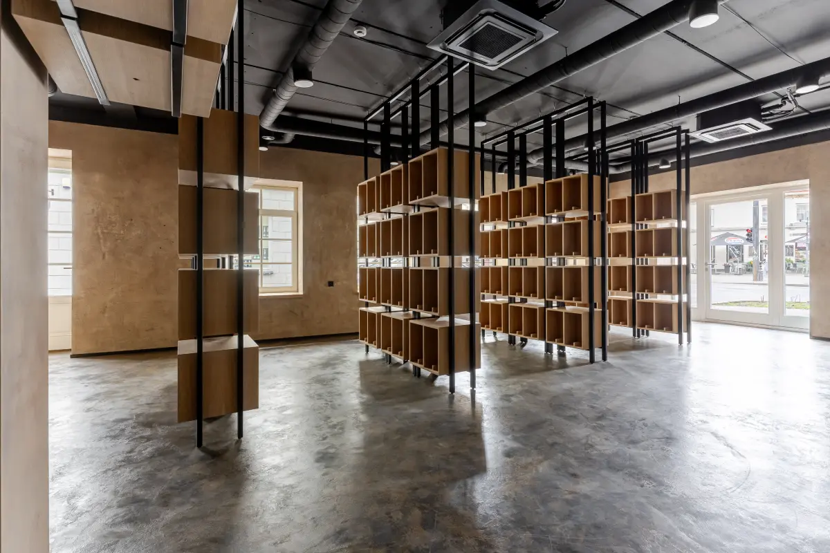 Tuščias modernus kambarys su medinėmis lentynomis, metalinėmis laikymo struktūromis ir dideliais langais, leidžiančiais patekti natūraliai šviesai.