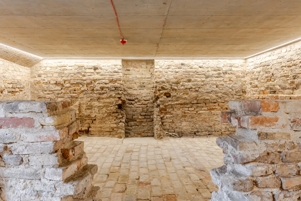 Istorinės plytų sienos ir grindys apšviestoje požeminėje patalpoje su betoniniu lubų įrenginiu.