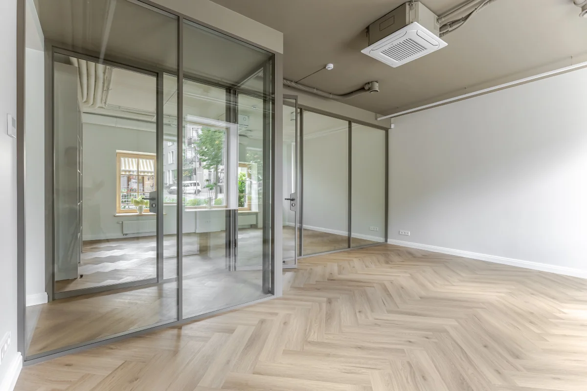 Šviesus tuščias kambarys su įstiklintomis pertvaromis, šviesios medinės grindys ir lubų vėdinimo sistema.