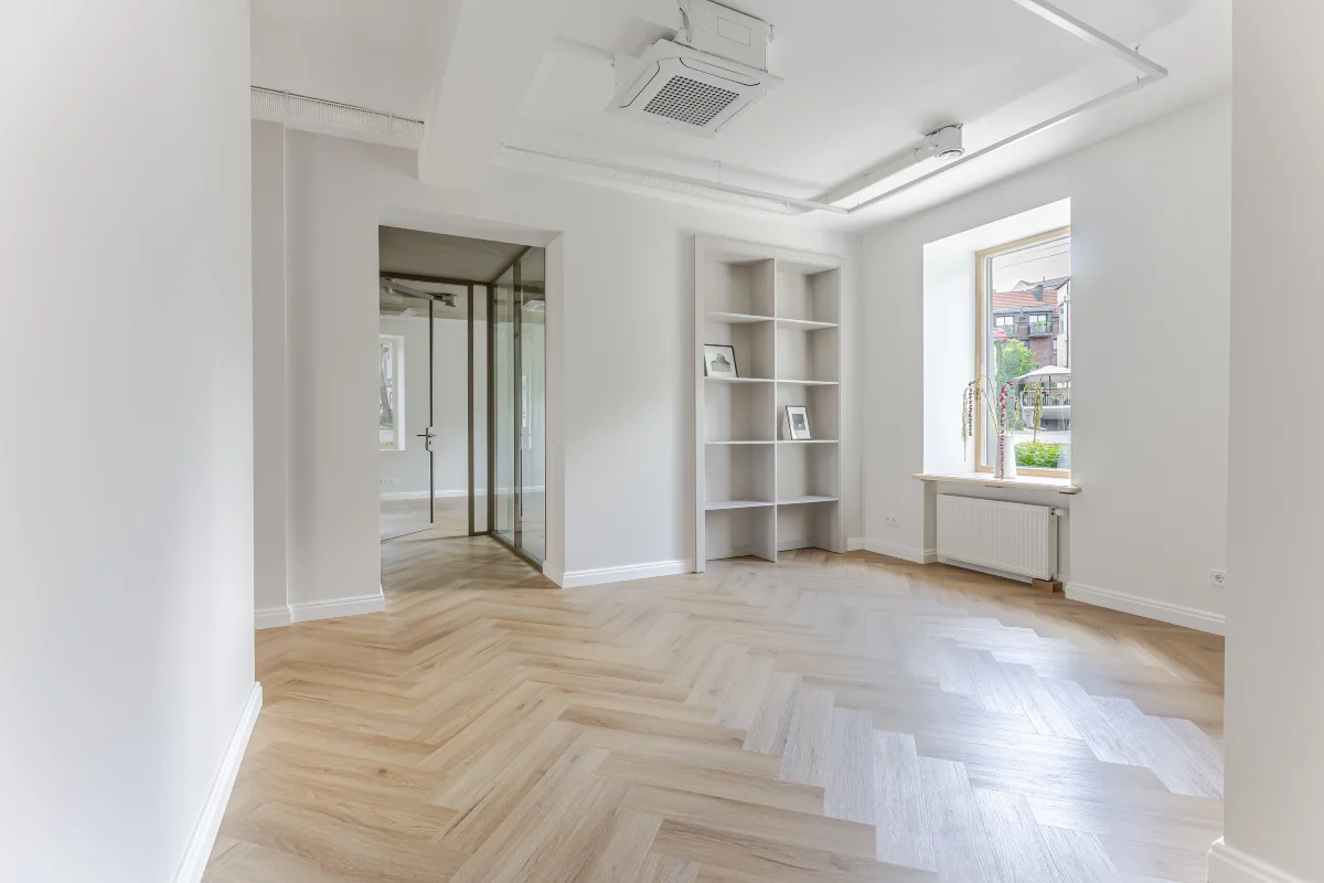 Šviesus tuščias kambarys su medinėmis grindimis, įmontuotomis lentynomis ir dideliu langų su vaizdu į miestą.