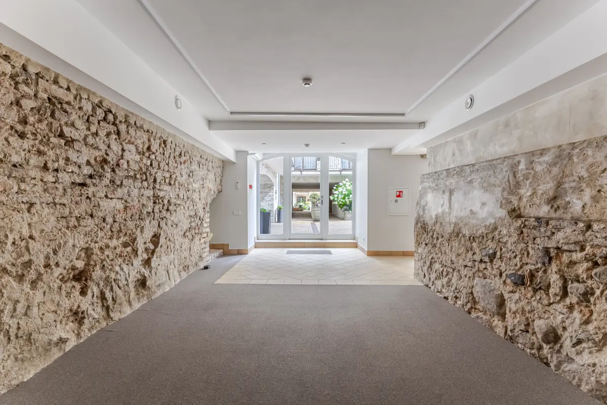 Įėjimas su plytų ir akmenų sienomis bei stiklinėmis durimis, vedančiomis į gėlių apželdintą kiemą.