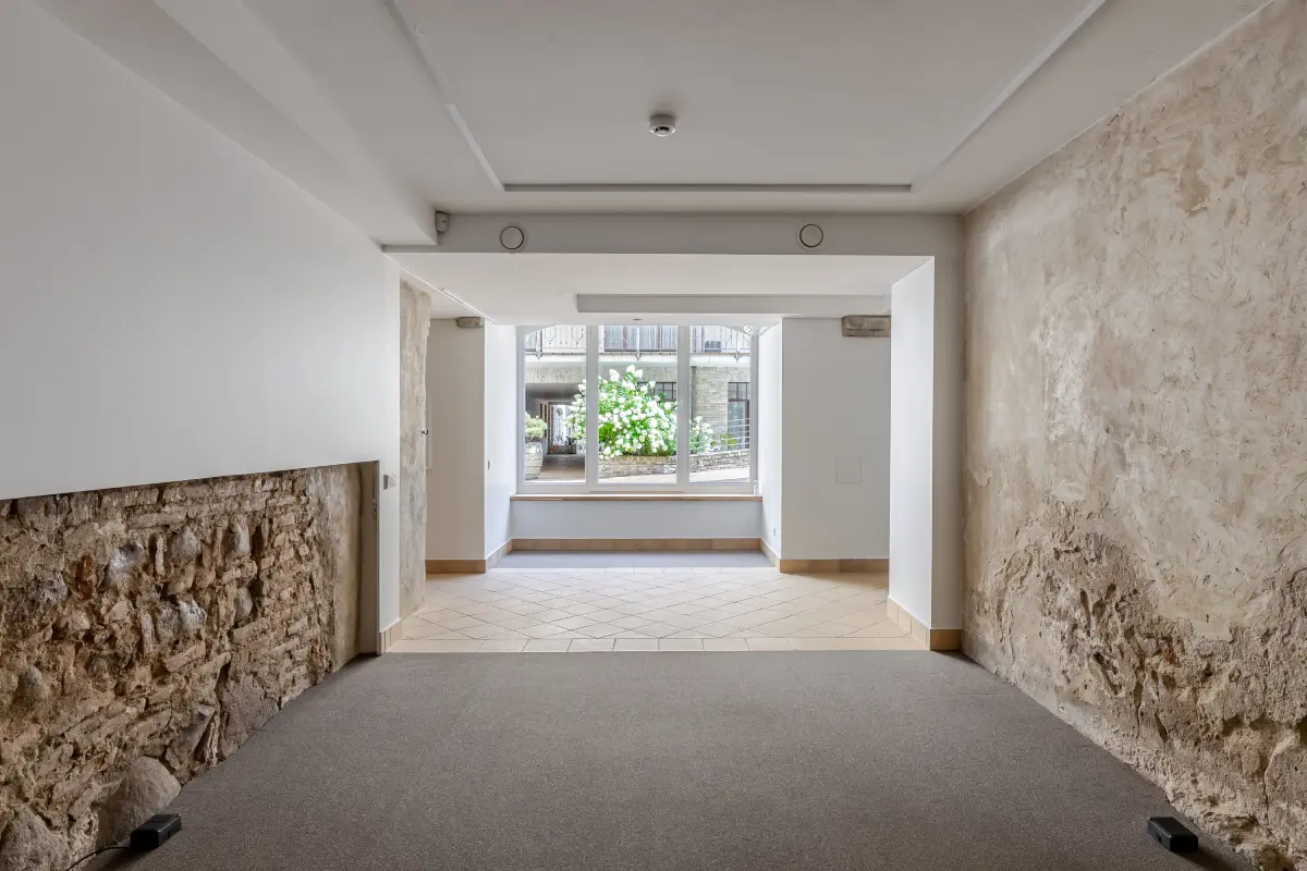 Tuščias kambarys su akmeninėmis sienomis ir dideliu langu, pro kurį matyti lauko gėlės.