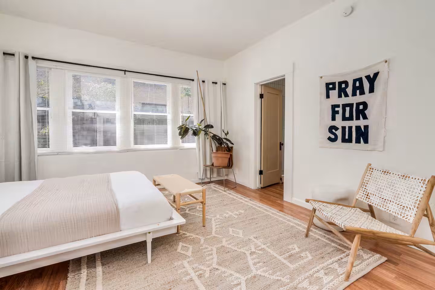 Bright bedroom with white walls, neutral bedding, woven bench, and ‘Pray for Sun’ wall art in a desert-inspired cottage.