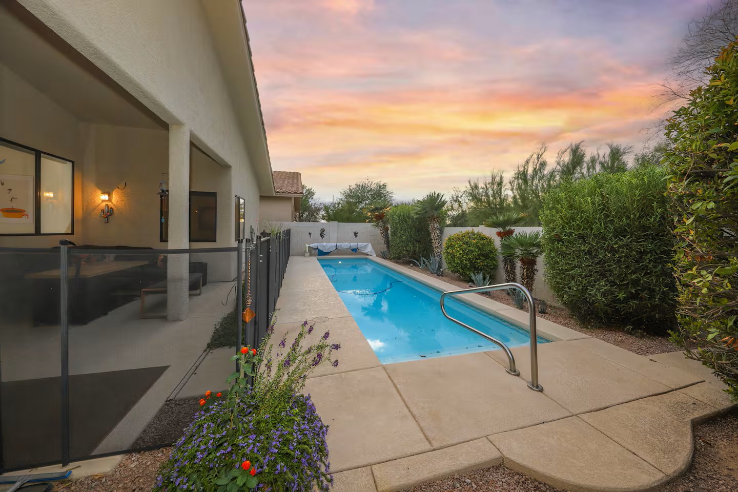 Backyard pool area at sunset featuring a long blue swimming pool, covered patio seating, and lush landscape at a Tucson vacation home.