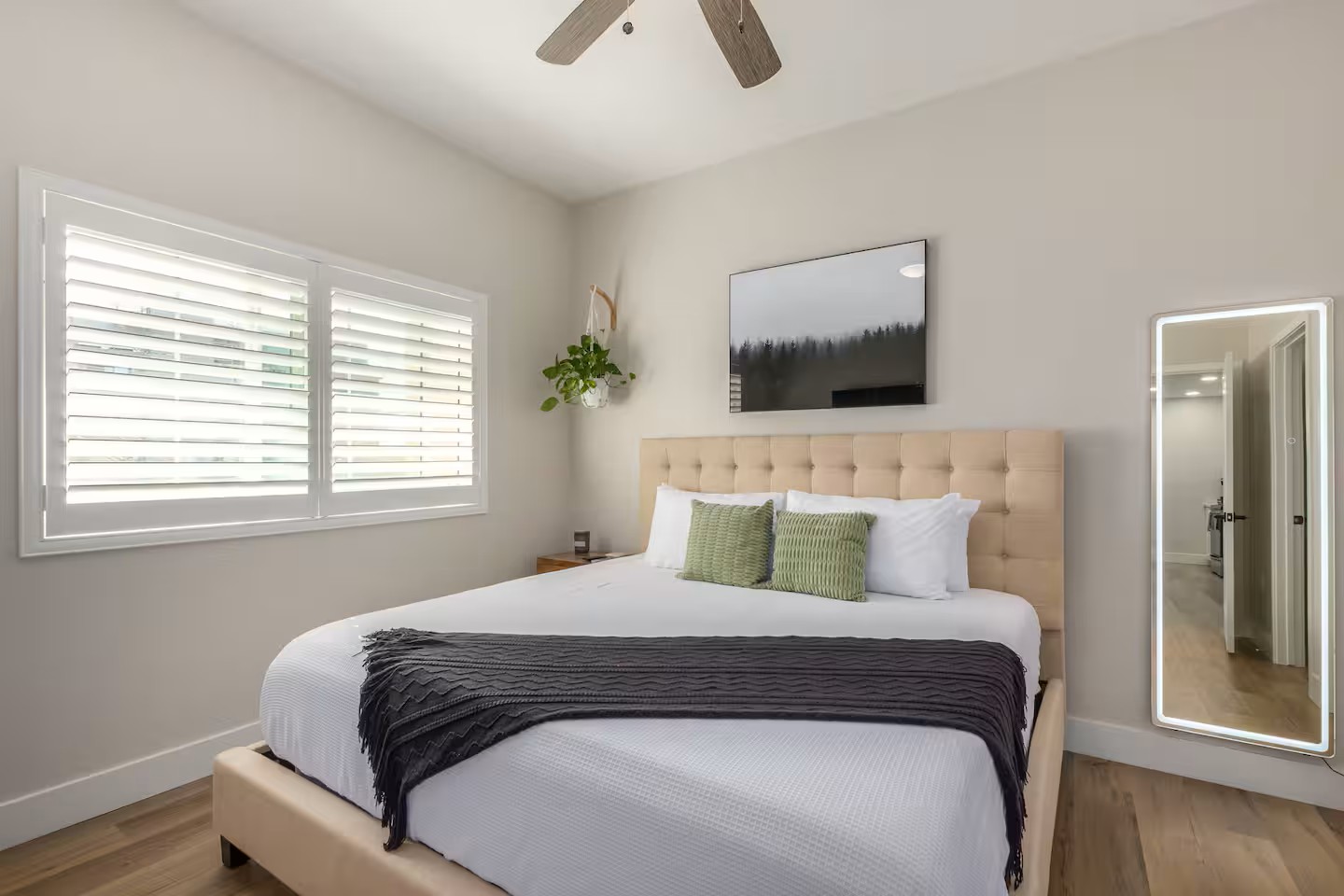 Bright modern bedroom with a tufted beige headboard, white bedding, green accents, and large windows with plantation shutters in a Tucson vacation rental.