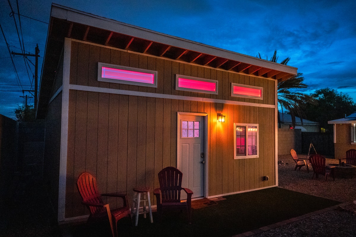 Modern tiny house exterior lit at night with warm accent lighting, vertical wood siding, and a cozy outdoor seating area.