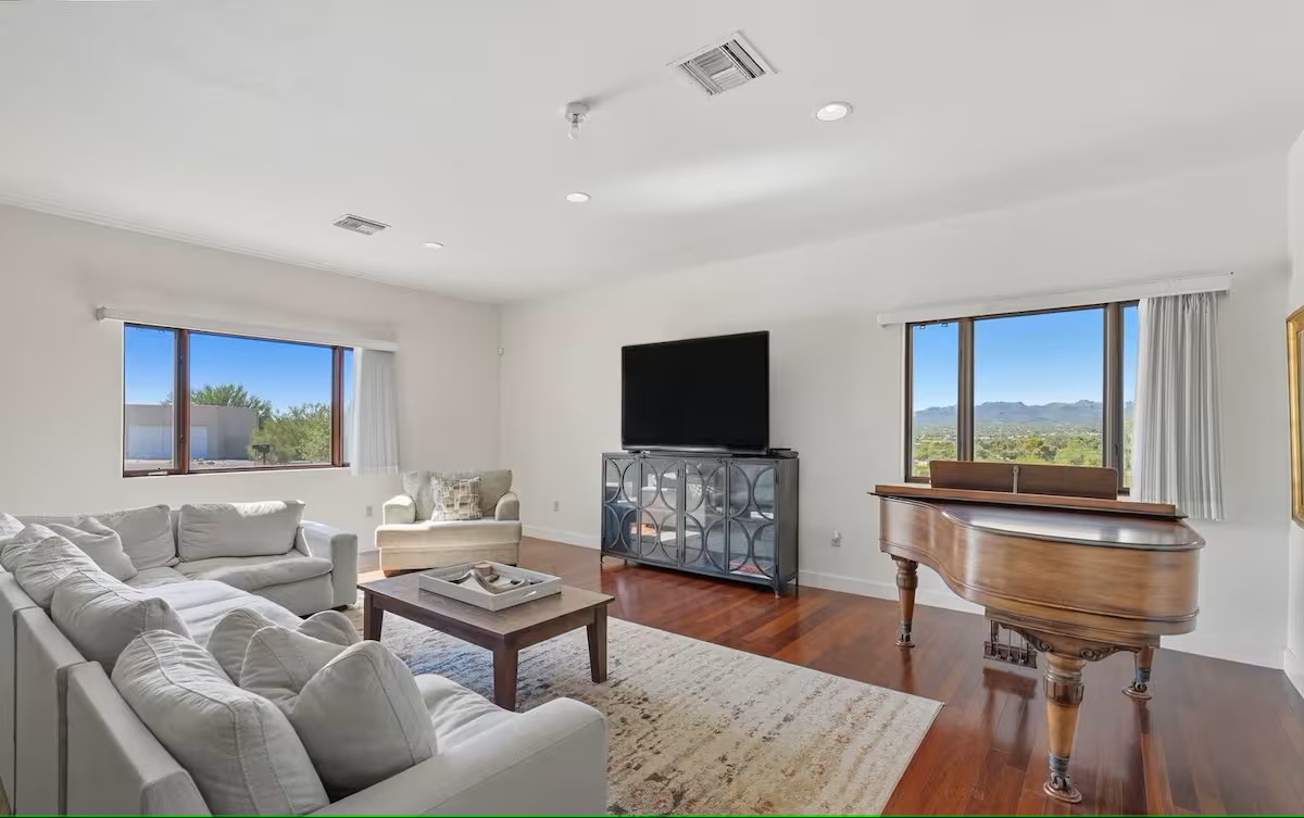 Spacious living room featuring large windows with desert views, light gray sofas, a grand piano, and warm wood flooring.